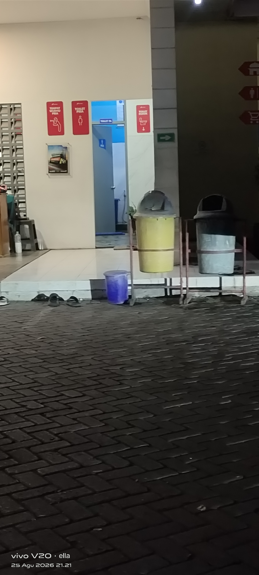 The image shows the exterior of a building with signs indicating a public restroom and a wudu area. The signs are in Indonesian and feature pictograms. To the left of the entrance, there are vertical ventilation grills. Inside the open doorway of the restroom, a blue toilet is visible. Outside the restroom, two large trash cans are placed on metal stands, with a smaller blue trash bin to the left. Several pairs of sandals are placed on the tiled area in front of the building. The ground is paved with dark brickwork. In the background, a darker, unlit area suggests the continuation of the building or an adjacent structure, with directional signs pointing to various facilities. The image appears to have been taken at night, as indicated by the lighting conditions and the timestamp at the bottom left corner which reads "25 Agu 2026 21.21". The location is identified as Tanjungsari, Indonesia.