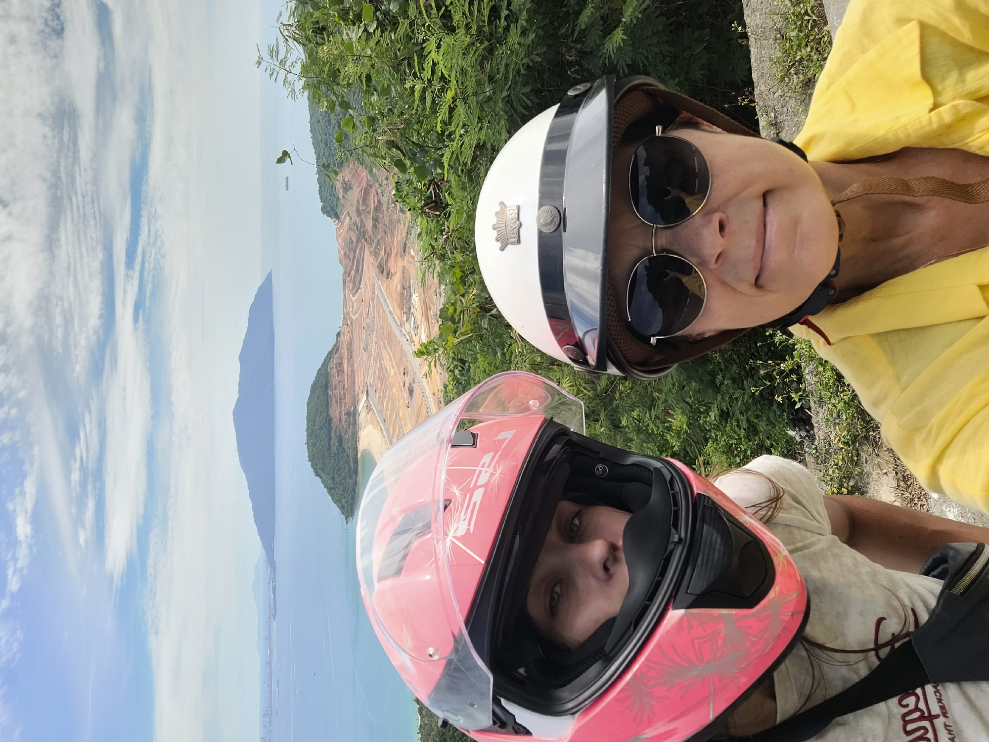 Two individuals are taking a selfie on a motorbike, with a scenic coastal and mountainous landscape in the background. The individual in the foreground on the left wears a pink motorcycle helmet with a clear visor and a white t-shirt. Their face is partially visible, showing their eyes. The individual on the right, also wearing a helmet, has a white and black design and wears round sunglasses and a bright yellow collared shirt. They are both positioned on what appears to be a road or lookout point overlooking a bay.
The background reveals a bay with calm blue water, a distant island or peninsula with a flattened summit, and a coastline featuring a sandy beach and a large construction site with cleared earth and roads. Mountains rise in the distance, framing the scene. The sky is partly cloudy with a bright, hazy atmosphere. The vegetation in the foreground consists of lush green bushes and trees.
The construction site in the background suggests development activity, potentially for a resort or infrastructure project. The location is likely along the Sơn Trà Peninsula in Đà Nẵng, Vietnam, known for its scenic views, beaches, and ongoing development. The presence of motorbikes and helmets indicates travel by motorcycle, a common mode of transportation in Vietnam. The overall scene captures a moment of adventure and appreciation for the natural beauty of the area, juxtaposed with signs of urbanization.