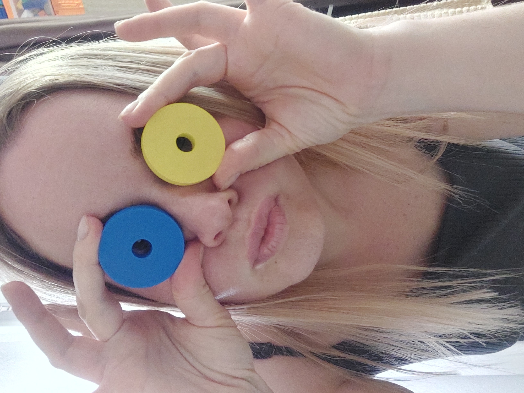 A close-up, diagonally oriented image captures a person, likely an adult with blonde hair, engaged in a playful activity indoors. The subject holds two colorful, circular objects – one bright yellow and one bright blue, both featuring a central hole – over their face, positioning them as if they were eyes or large spectacles. The yellow disc is held over the person's right eye area by their right hand, while the blue disc is held over the left eye area by their left hand.
The person's hands are prominent, with fingers gently gripping the discs. Parts of the subject's face are visible, including the nose and a partially open mouth, suggesting a relaxed or whimsical expression. Blonde hair cascades around the face, contributing to the overall composition. The background is softly blurred and indistinct, indicating an indoor setting, possibly a home, with hints of furniture or walls. The lighting is soft and even, consistent with indoor conditions. There are no specific cues to determine the exact time of day or external weather, nor are there any visible texts. The image conveys a lighthearted and creative moment.