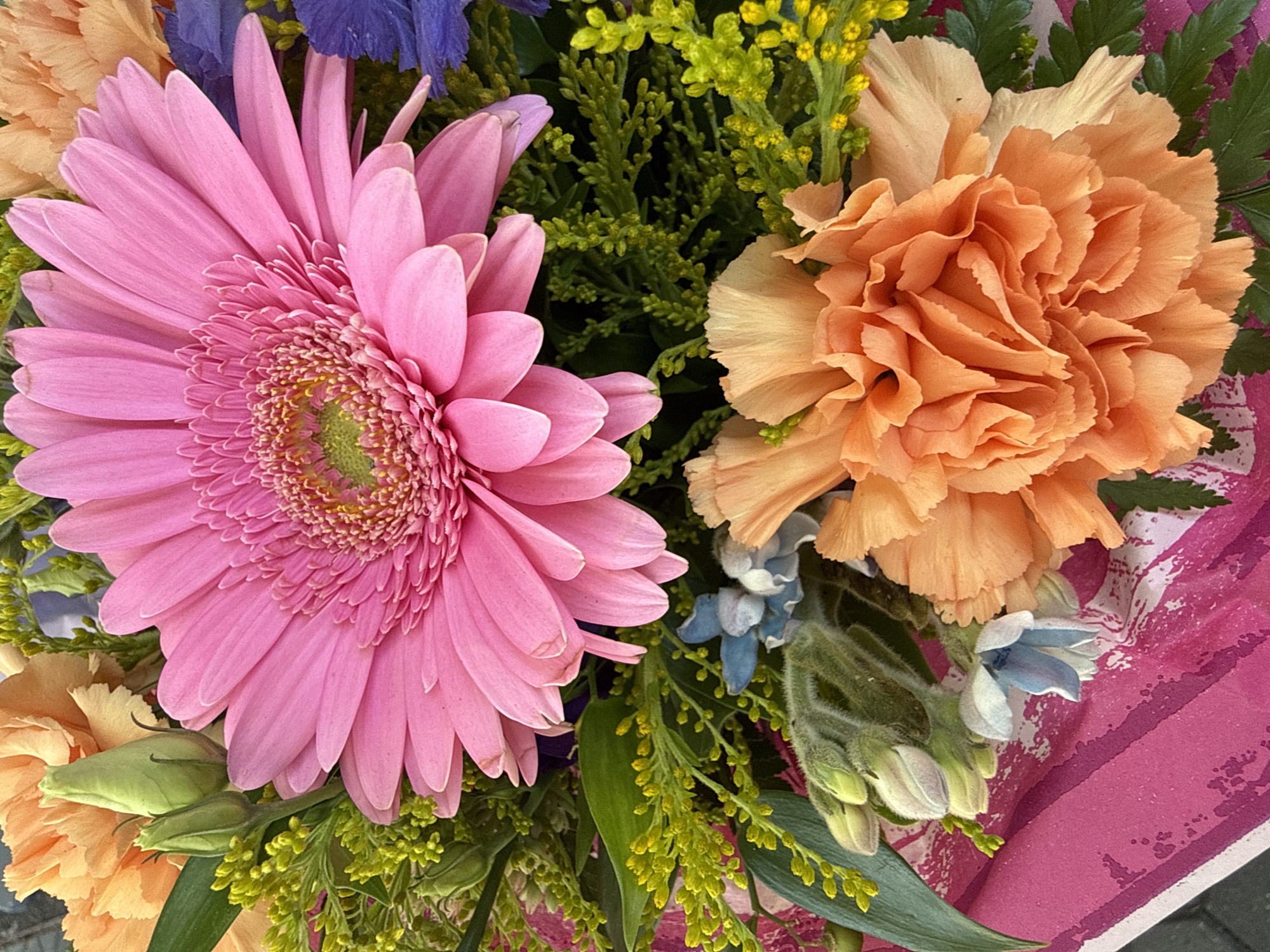 This is a close-up, overhead shot of a colorful flower arrangement. The bouquet features a large, vibrant pink Gerbera daisy as the prominent centerpiece. Surrounding the Gerbera are several peach or orange carnations, small clusters of delicate blue flowers, and sprigs of green filler flowers and foliage. The arrangement is presented against a background of bright pink wrapping paper with a textured, abstract pattern. No discernible city or location information is present in the image.