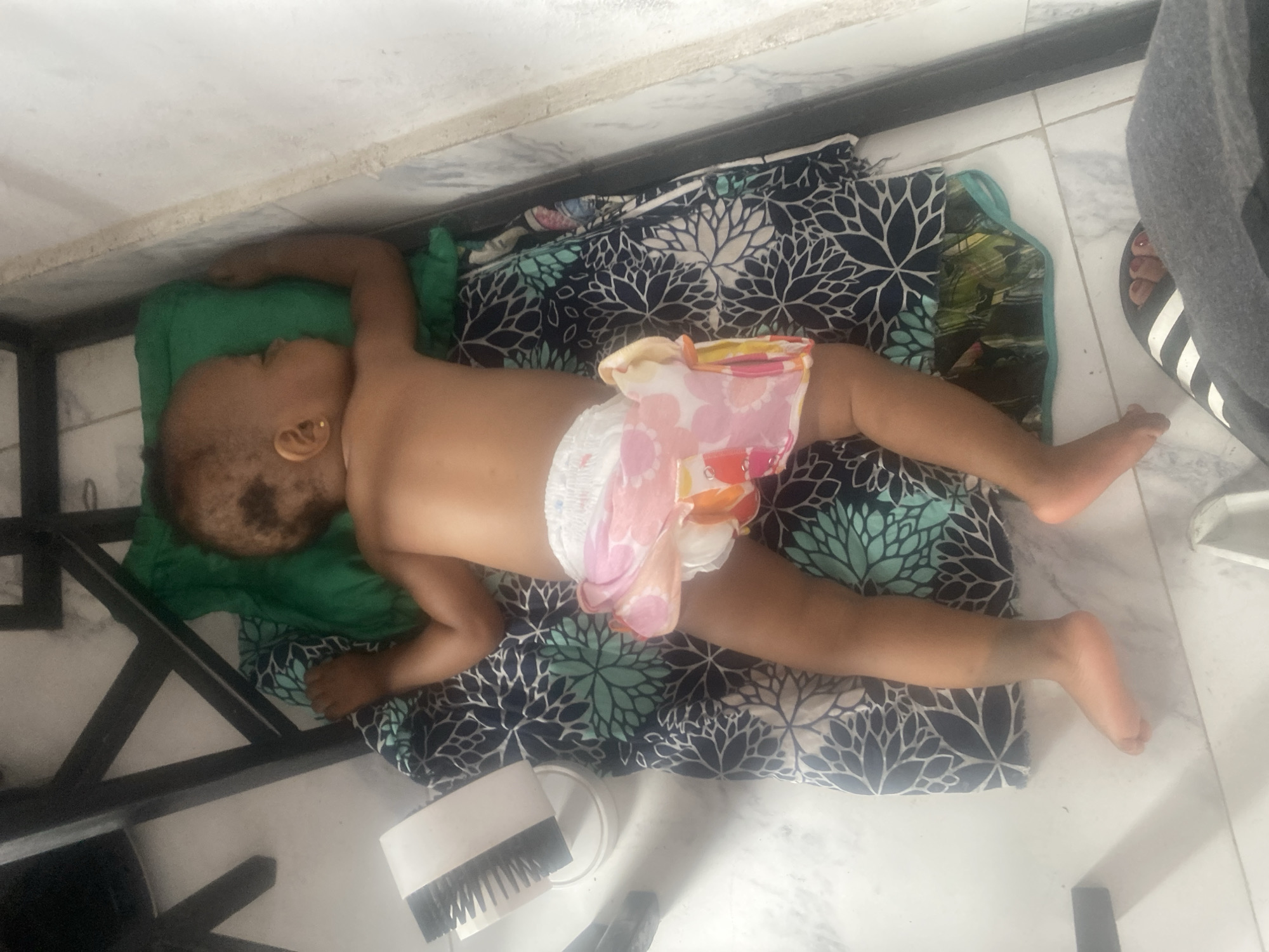 An overhead shot shows a baby or young child sleeping soundly on its back on the floor. The child has light brown skin, short dark hair, and is wearing a white diaper with pink and orange floral patterns. A small gold stud earring is visible in the child's right ear. The child is resting its head on a green pillow or cushion and is lying on a patterned mat or blanket featuring dark blue fabric with light blue and white leaf or flower designs.
The setting appears to be indoors, likely in a home in Lekki, Nigeria, given the provided context. The floor is covered with white or light grey tiles, indicating an indoor environment. A dark metal frame structure, possibly part of a bed frame or other furniture, is partially visible in the upper left and lower left of the frame. To the right, part of an adult's lower leg and foot, wearing a black and white striped slide sandal, can be seen, along with grey fabric, possibly trousers or shorts. Near the child's lower body, a white object with black bristles, resembling a small brush, and a white circular object (possibly a lid) are on the tiled floor. The overall impression is one of peaceful rest. There is no visible text in the image.