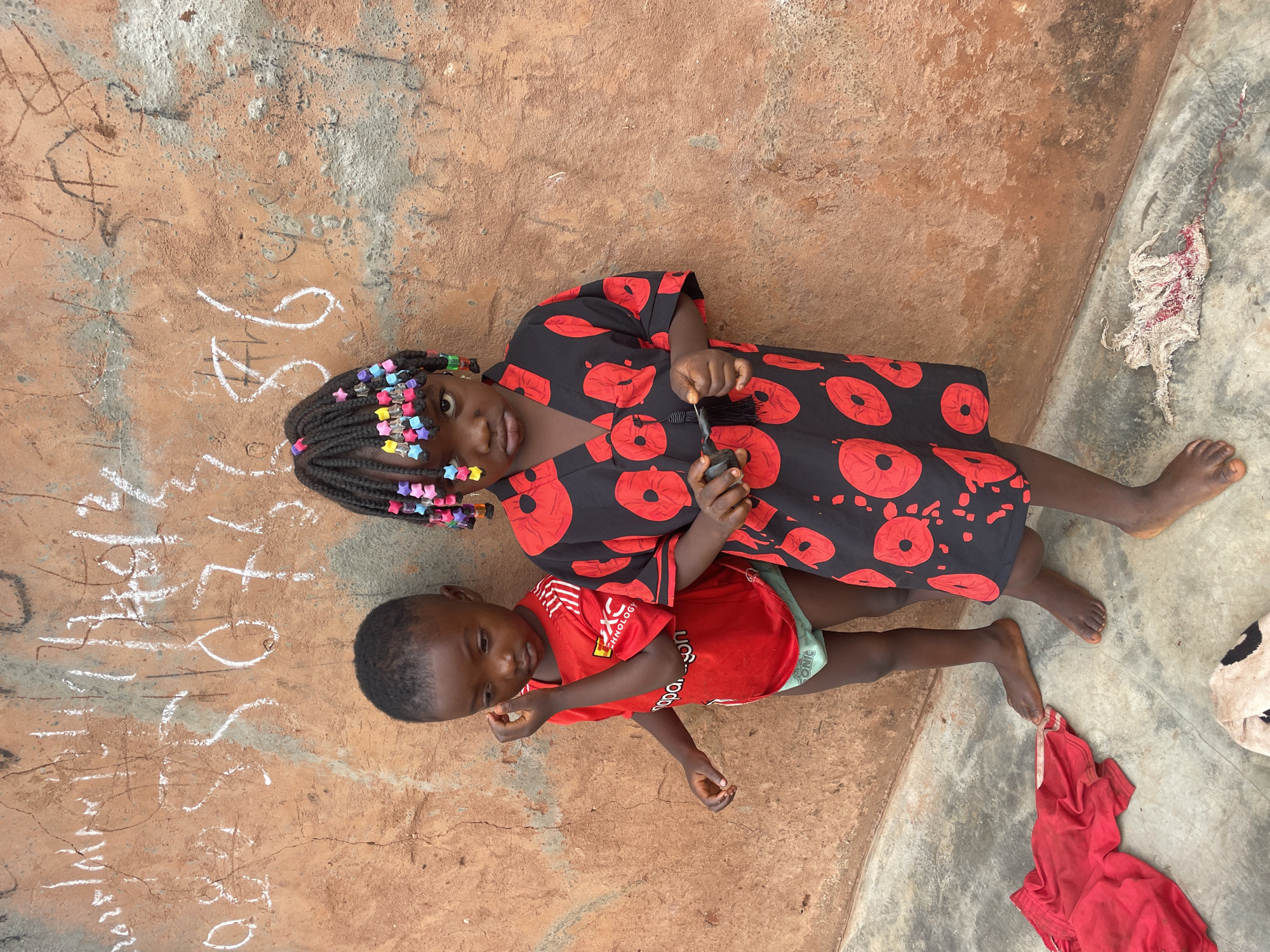 Two children, likely siblings, are on the ground in Ogbe, Nigeria. The older child, a girl with braided hair decorated with colorful beads, wears a red and black patterned dress. She is holding a small, dark object. The younger child, a boy, wears a red t-shirt. The children are on a surface that is a combination of dirt and concrete. There is graffiti or writing on the dirt section. A red t-shirt is also on the ground near the concrete section.