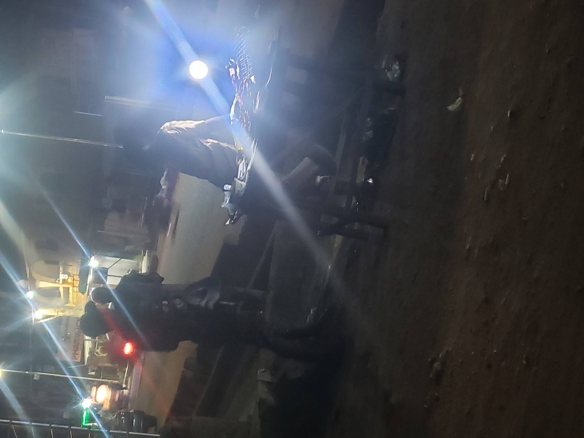 A dark image shows a night scene in Ibadan, Nigeria. A person is sitting on a motorcycle under a bright street light. A traffic light shows red and green, with the sign "PHARMACY" visible nearby. The area appears to be a street with buildings and other street lights in the background. The ground is dark, suggesting either a dirt or paved surface.