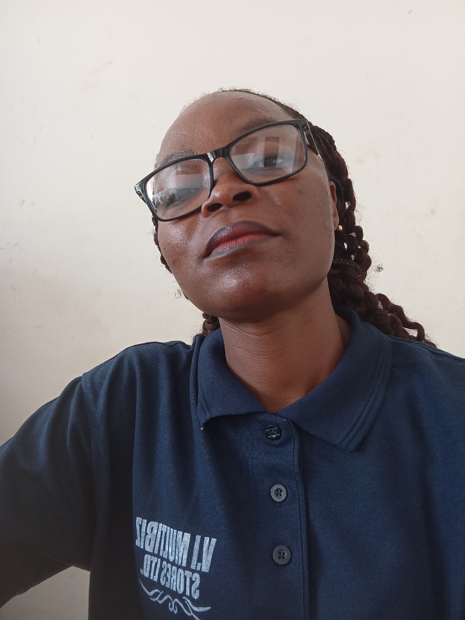 A medium shot photo shows a person with dark skin wearing glasses and a dark blue collared shirt. The person is looking upwards and slightly to the left of the camera. Their hair is braided and visible behind their ears. The background is a plain, light-colored wall. A logo is visible on the person's shirt, but it is partially obscured and difficult to read.