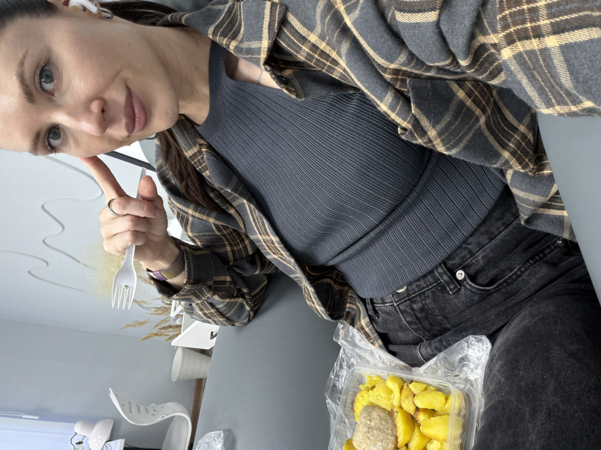 A woman is holding a plastic fork and giving a thumbs-up while seated with a container of potatoes and what appears to be a meat patty beside her. She is wearing a dark gray ribbed top, black jeans, and a gray and brown plaid shirt. The location is possibly Perm, Russia based on the woman's appearance, but there are no definitive cues in the image to confirm.