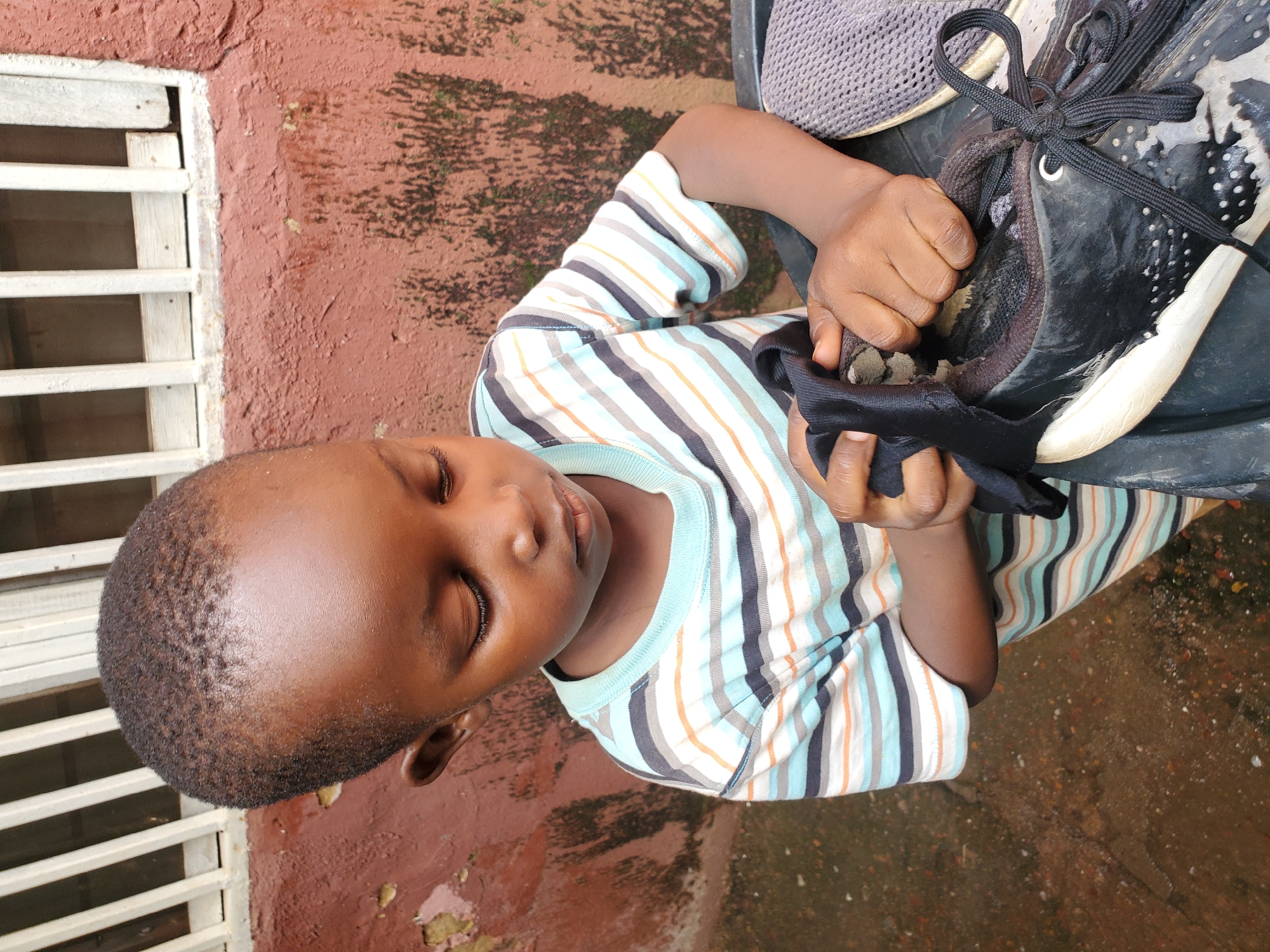 A close-up, high-angle shot shows a young Black child, viewed from the head down, wearing a horizontally striped t-shirt in shades of blue, white, gray, and orange. The child's eyes are closed, and they appear to be resting or asleep. Their right hand is holding a black shoe, the laces of which are prominent. The child is positioned in an outdoor setting, with a reddish-brown, textured wall on the left, partially covered by a white grate. The ground appears to be a mix of dirt and scattered pebbles. The overall lighting suggests it is daytime. The context of Jos, Nigeria is provided, though not visually evident in the image itself.