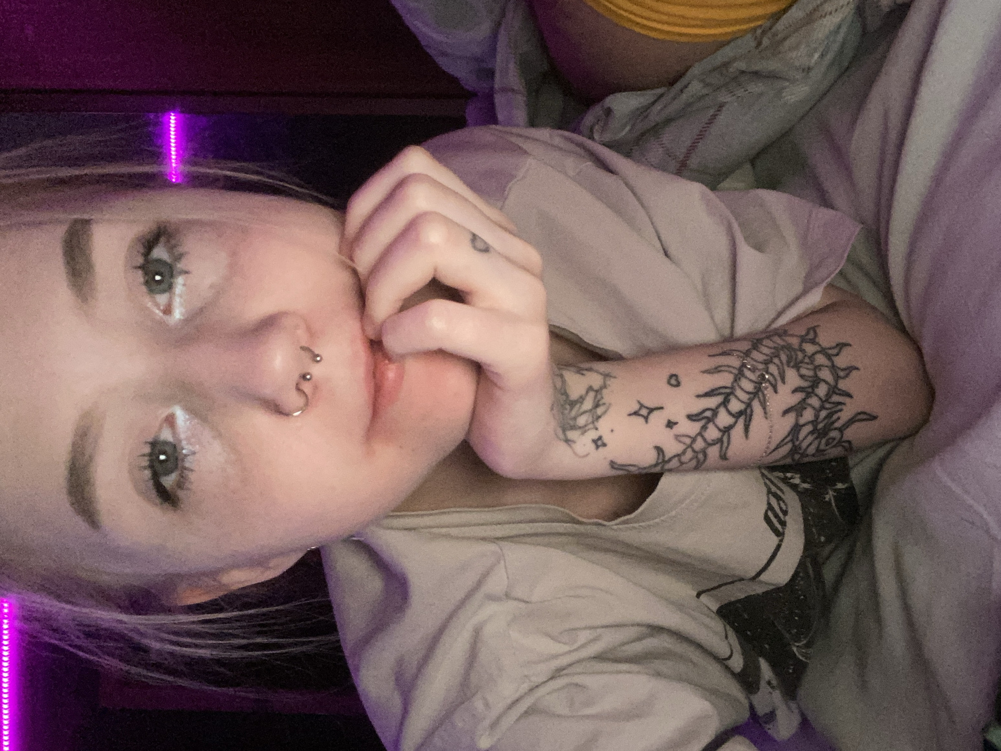 The scene depicts a close-up selfie of a young, light-skinned individual, likely female, situated indoors in Izhevsk, Russia. The subject has blonde hair, green eyes accentuated with white eyeliner on the lower lash line, natural eyebrows, a septum piercing (gold-toned hoop), and a bridge piercing (silver-toned barbell). The individual is shown resting their chin on the curled fingers of their right hand.
The subject is wearing a light beige or light grey short-sleeved t-shirt. Tattoos are visible on the left forearm, featuring a detailed arthropod-like creature (resembling a centipede) surrounded by small star motifs. A small, dark heart-shaped tattoo is present on the ring finger of the right hand.
The environment is dimly lit, with a prominent strip of magenta/purple LED lighting visible along the top left edge of the frame, casting a purple glow on the subject's hair and surrounding areas. The background is dark and indistinct. The individual appears to be reclining on soft bedding, with shades of light grey and purple fabrics visible, along with a portion of a yellow-orange fabric on the far right. No other individuals or specific objects are clearly discernible.
