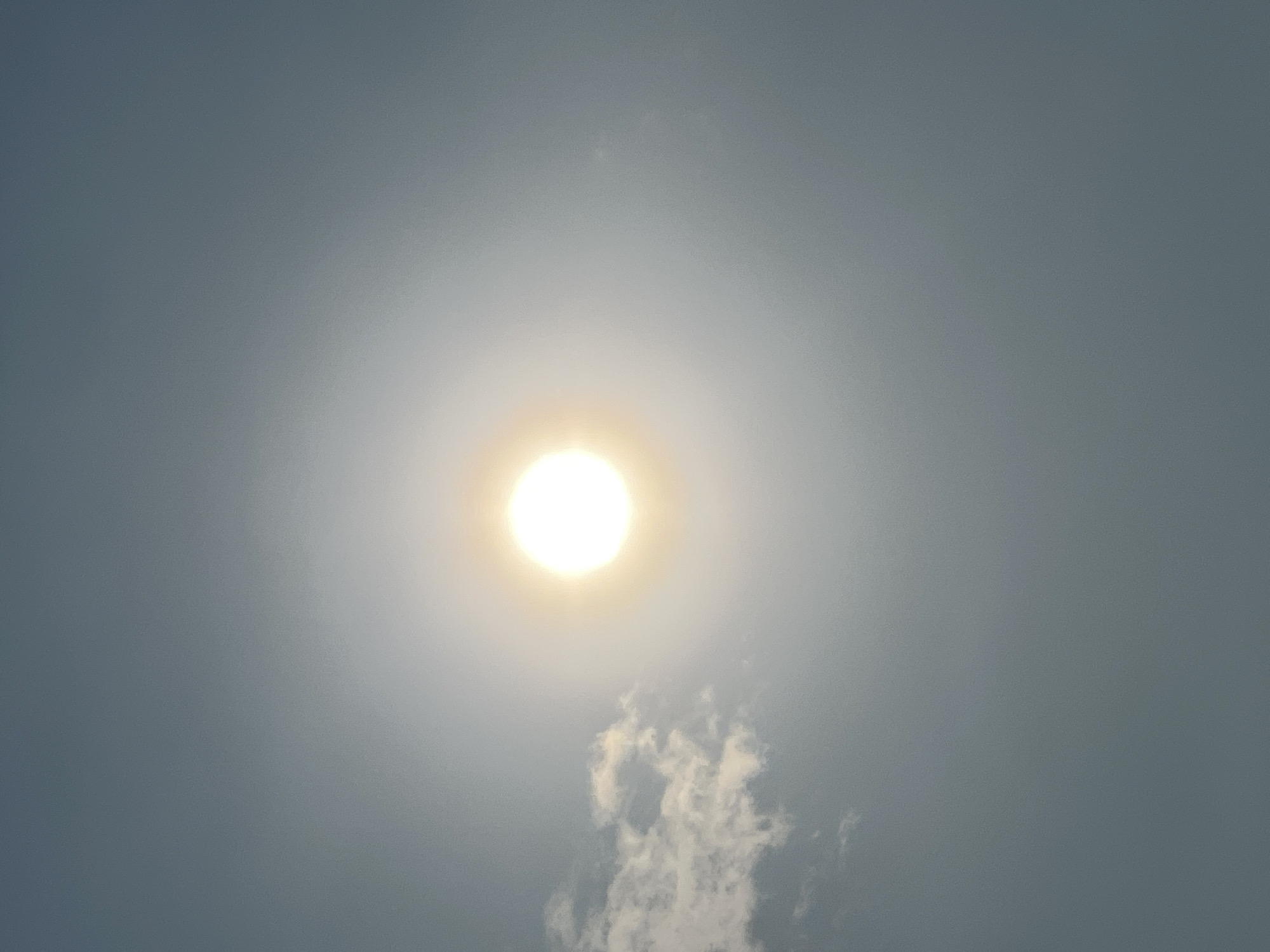 The image depicts a bright sun partially obscured by a hazy sky. A small cluster of clouds is visible to the left of the sun. The overall scene suggests a bright, possibly warm day, with the sun creating a luminous halo effect. The sky is a gradient of light blue to grey, indicating the presence of atmospheric haze or light cloud cover. There are no people or discernible structures visible in the frame. The location is identified as Abuja, Nigeria, though no specific landmarks or contextual cues within the image confirm this.