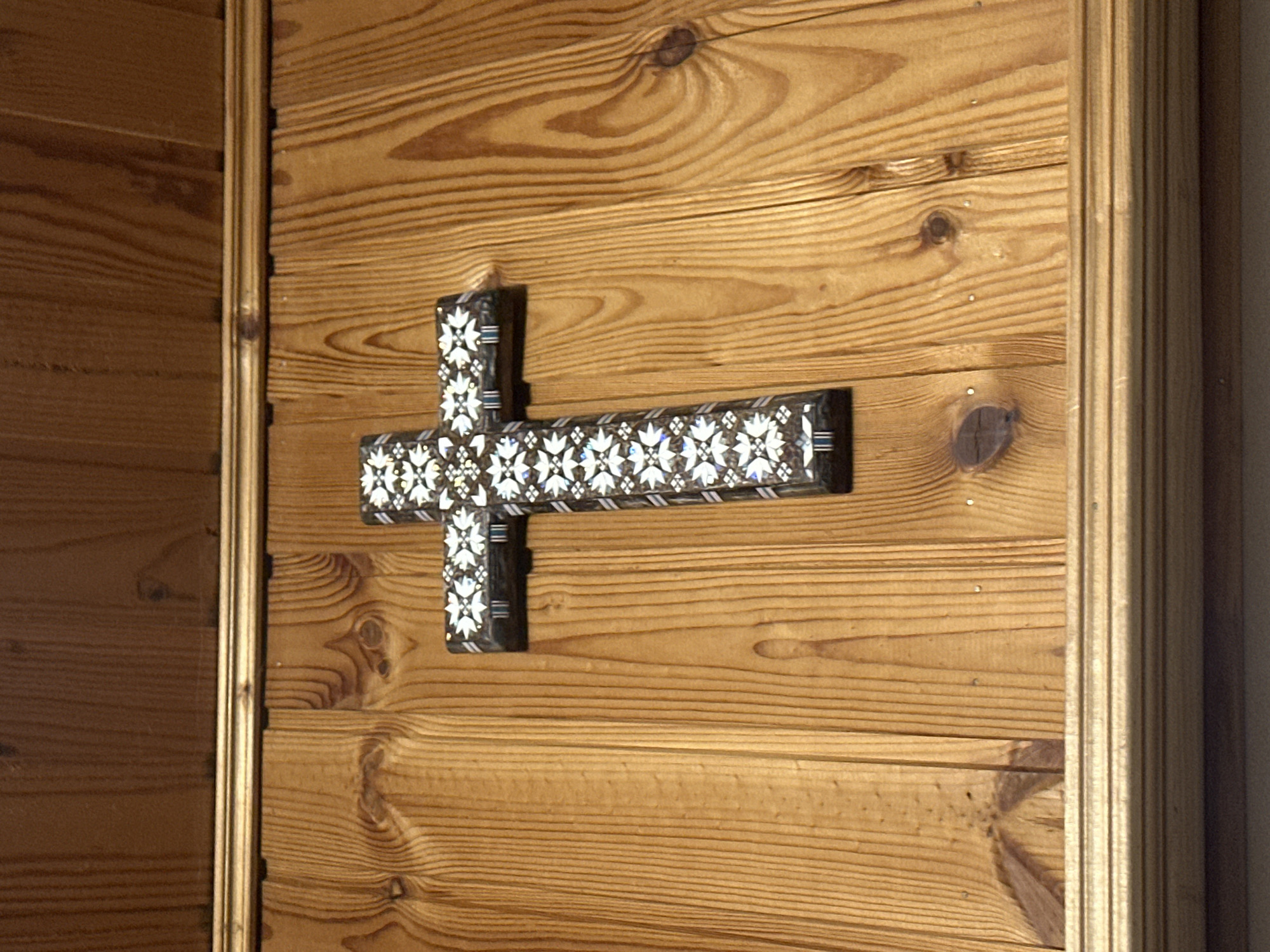 The image shows a wooden door with a decorative cross attached to it. The cross is dark in color and adorned with a pattern of white floral shapes. The door itself is made of light brown wood with a distinct grain pattern. The image was taken at Kurlutovka, Russia.