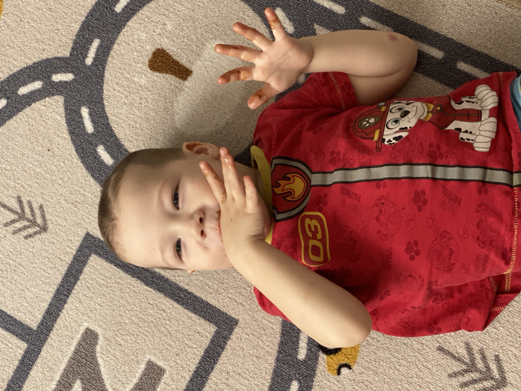 A young child is captured in a close-up, overhead shot, lying on their back on a light-colored rug. The child, who appears to be a toddler or preschooler, has short, light brown hair and is looking directly at the camera with a slight, playful smile. Their eyes are dark and focused.
Both of the child's hands are raised, positioned near their head and face. There is a dark, brown, sticky substance covering the child's fingers and also visible around their mouth, suggesting they may have recently eaten something like chocolate or mud. A small, reddish mark or abrasion is visible on their right arm, near the shoulder.
The child is wearing a red, long-sleeved shirt that prominently features characters from the children's show, Paw Patrol. On the chest of the shirt, a large graphic of Marshall, the dalmatian firefighter dog, is visible. Below Marshall, there's a yellow number "03" in a block font, and to the left, a yellow flame emblem. The red fabric of the shirt has a subtle, repeating pattern of "PAW PATROL" text, paw prints, and other Paw Patrol characters.
The rug beneath the child is beige or light grey with various patterns. One part features a grey road with white dashed lines, suggesting a play mat. Another section shows geometric patterns, including grey and brown arrow-like shapes. The overall setting appears to be indoors, likely in a home environment. The messy hands and face add a candid and typical childhood moment to the image.