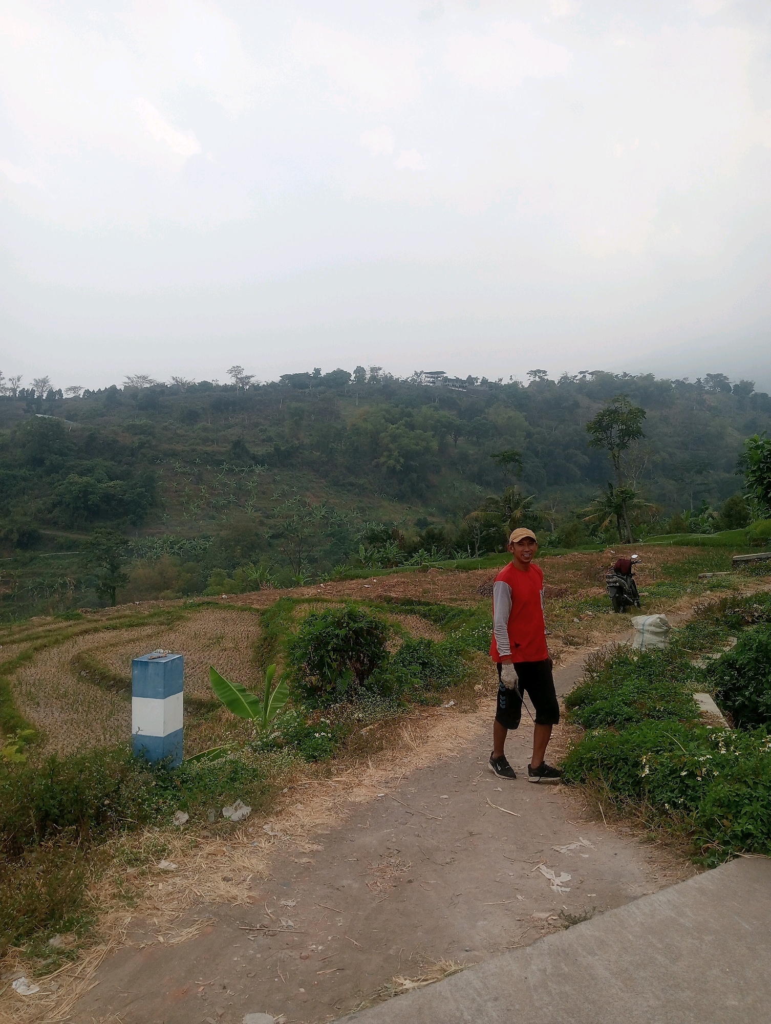 A young man wearing a red shirt and black shorts stands on a dirt path in a rural area. The path is surrounded by dry grass and green bushes. In the background, there are terraced fields and lush green hills covered in trees. The sky is overcast. A blue and white marker post is visible to the left of the path, and a motorcycle is parked behind the man.