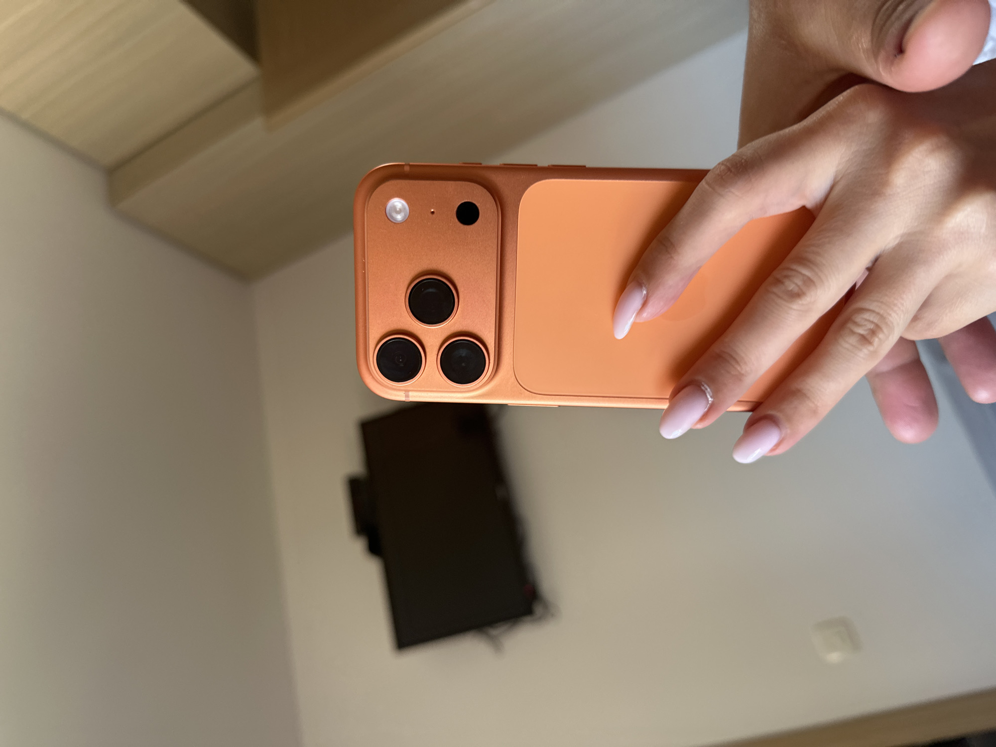 The image shows a person holding an orange smartphone. The smartphone has a camera module with three lenses and a flash. In the background, a television is mounted on the wall of a room. The room appears to be a bedroom or a hotel room, with a wardrobe visible on the right side of the image. The overall setting suggests an indoor environment, likely a private residence or accommodation. There is no visible indication of the specific location within Budva, Montenegro.