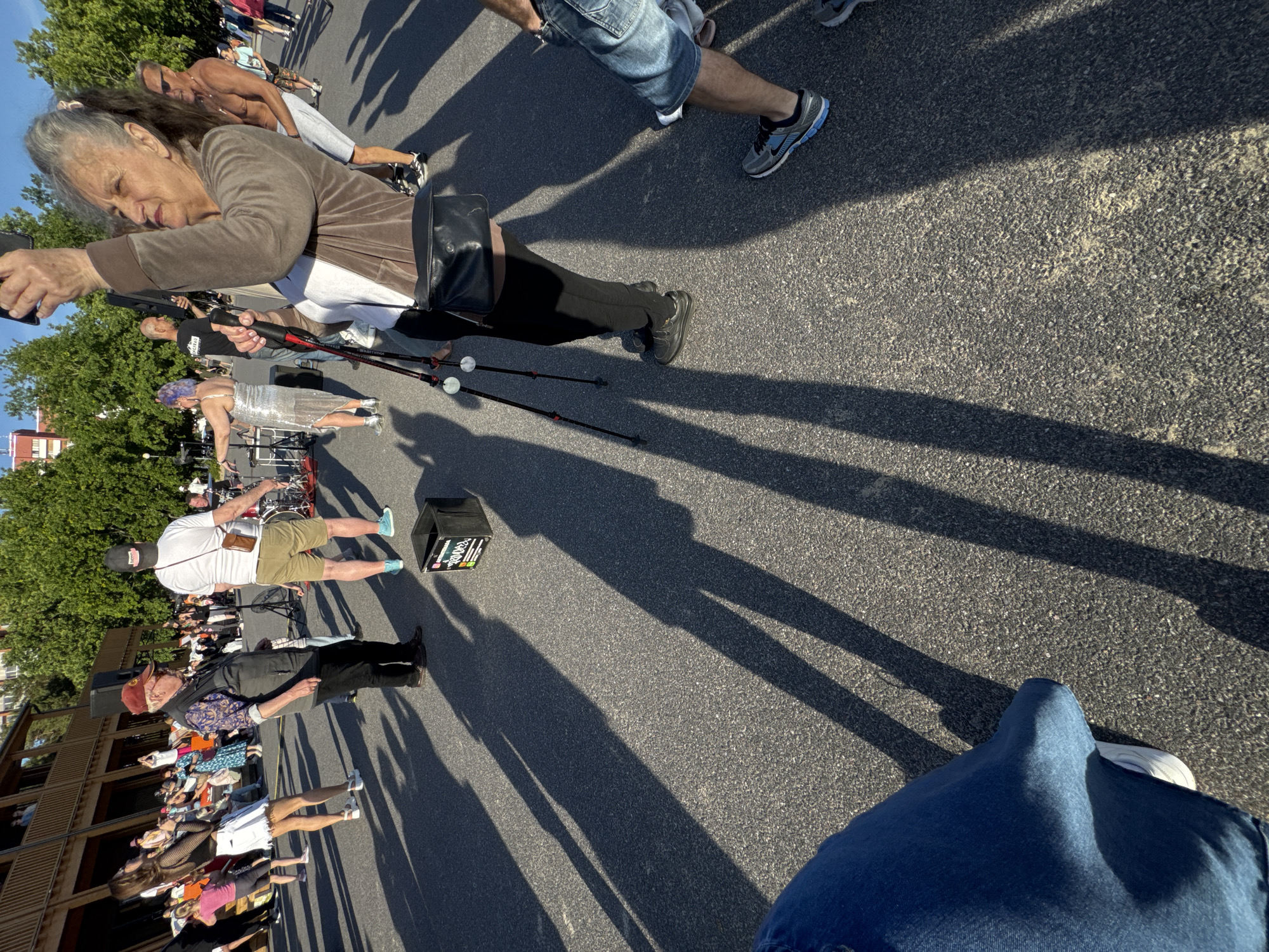 In the city of Tallinn, Estonia, a group of people are gathered outdoors on a paved area, likely a plaza or street, under a clear sky. The scene is bathed in warm sunlight, casting long shadows across the asphalt. In the foreground, a person wearing blue jeans is partially visible, their legs and feet facing towards the right side of the frame. To their left, a woman with gray hair pulled back in a ponytail is captured in a close-up, holding up a smartphone to take a picture or video. She is wearing a brown jacket over a white shirt and black pants, and has a black crossbody bag. She is holding two black hiking poles with red accents.
Behind her, a man with sunglasses and a bare chest, wearing light gray shorts, stands facing towards the left. Further into the scene, several individuals are visible. A man in a red cap and a patterned shirt, wearing a grey vest and dark pants, is looking to his left. To his right, a person in a white t-shirt and khaki shorts is facing away from the camera. A woman with purple hair, wearing a shimmering silver dress, is positioned further back, appearing to be dancing or posing. A drum set is set up to the right of the frame. In the background, a wooden building with multiple levels and balconies is visible, with trees and other structures beyond. Various other people are spread throughout the area, some standing and observing, others appearing to be in conversation or engaged in activities. A black plastic bin with stickers on it sits on the ground in the mid-ground.