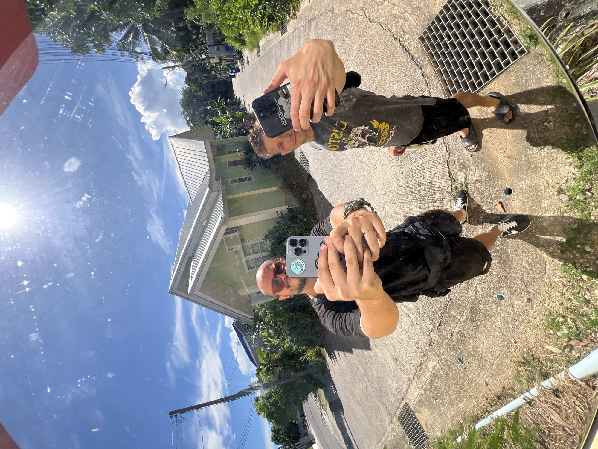 In a wide-angle shot taken outdoors in a residential area, two men are seen taking a selfie in what appears to be a reflective surface, possibly a car mirror. The man on the left, wearing sunglasses, a black t-shirt, and black shorts, holds a gray smartphone with a blue wave sticker. The man on the right, wearing a dark grey t-shirt with yellow lettering and black shorts, holds a black smartphone. They are standing on a paved surface next to a road with houses and lush vegetation lining the sides. The sky above is bright blue with scattered white clouds, and the sun is high in the sky, causing lens flare. The overall scene depicts a casual outdoor setting with a focus on the two individuals capturing their own image.