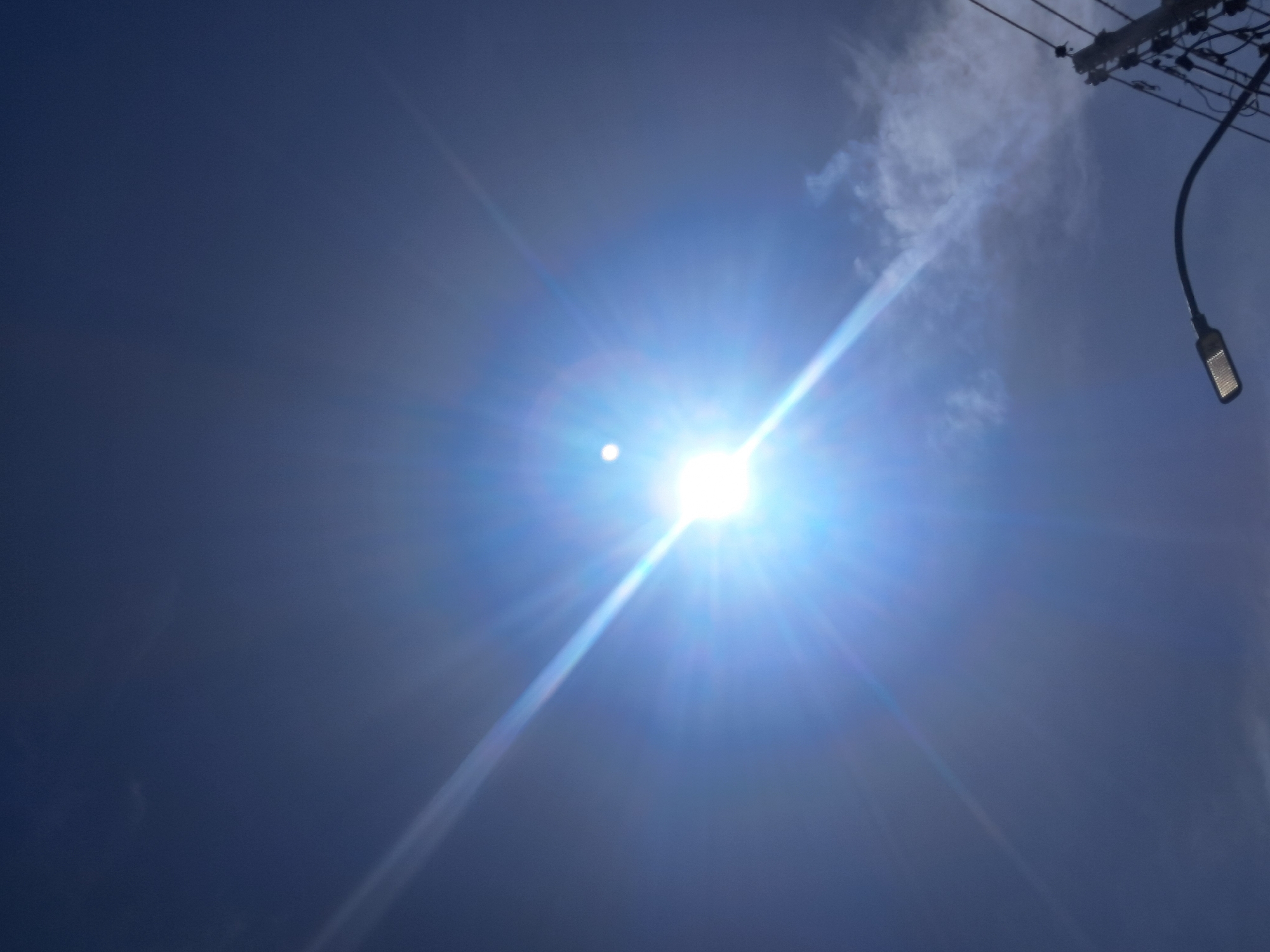 The image shows a bright sun in a dark blue sky with a small cloud cluster to the upper right. A street light and power lines are visible in the upper right corner. There are no people or actions visible in the frame. The location is specified as Juan Griego, Venezuela.