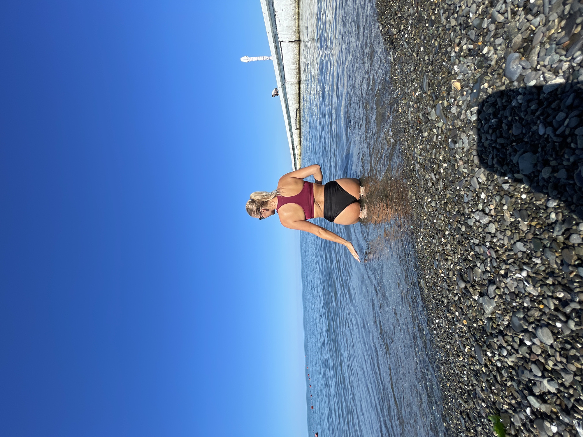 A blonde woman with a ponytail, wearing a maroon and black bikini, sits in the shallow water of the Black Sea. Her back is to the camera, and she is looking out at the water. Her left hand is resting on the water's surface. The water is clear and blue, with small waves lapping at the pebbly shore. In the background, a concrete pier extends into the sea, with a few figures visible on it. The sky is a clear, deep blue. The scene appears to be a typical summer day at the beach in Sochi, Russia.