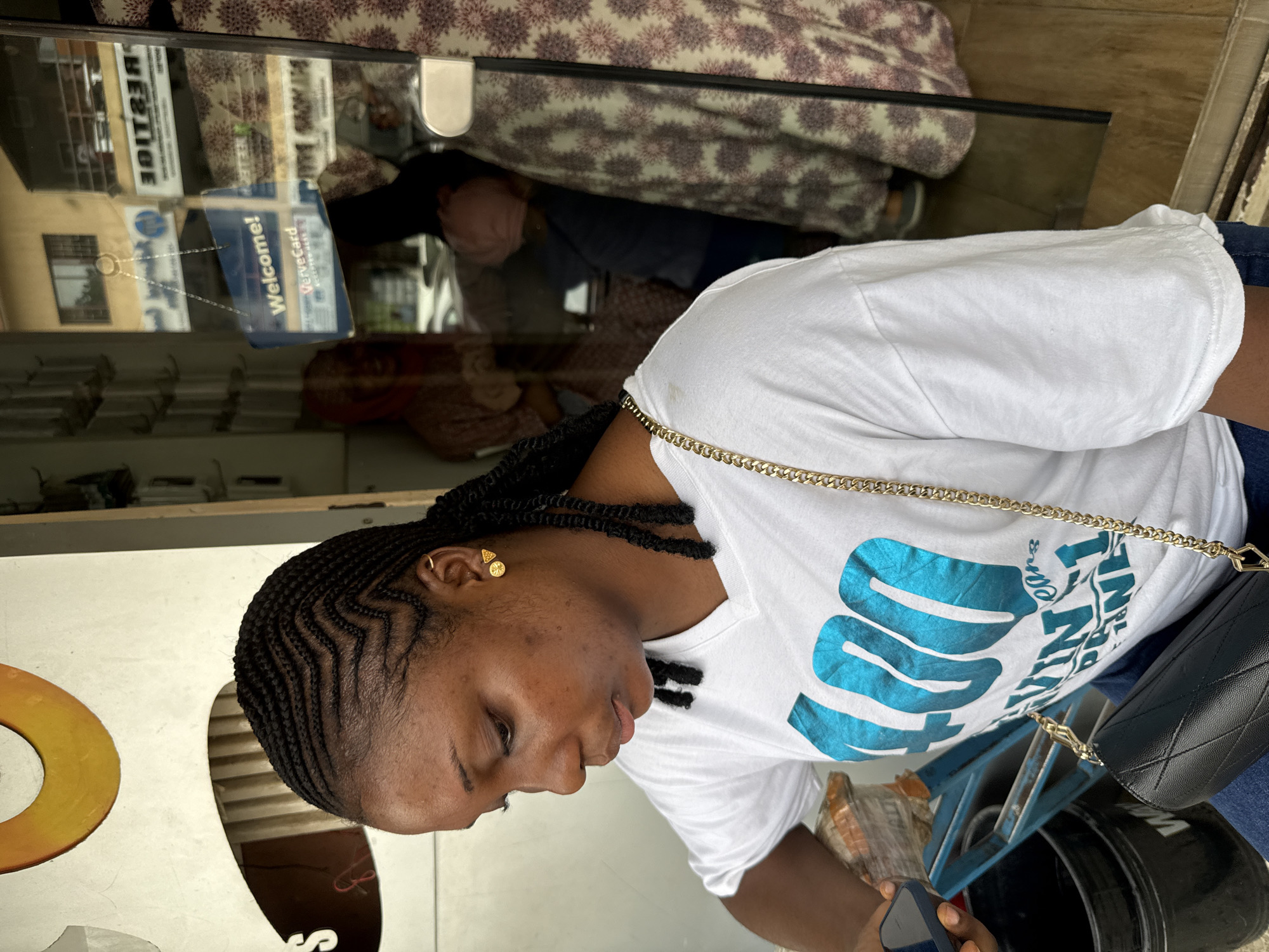 A young woman is the main subject of the image. She has dark hair braided tightly to her scalp. She's wearing a white t-shirt with blue lettering and a gold chain. Her right ear has a gold earring. She appears to be looking at a smartphone in her hands. A black quilted purse hangs from her shoulder.
The background includes a glass storefront window with reflections of people and buildings. In the reflection, a sign is visible that reads "Vervetcard, Welcome!". The building reflected in the window shows "Prestige" is written on it. The image appears to have been taken outdoors in daylight. I do not have sufficient information to identify the precise location as Abuja, Nigeria.