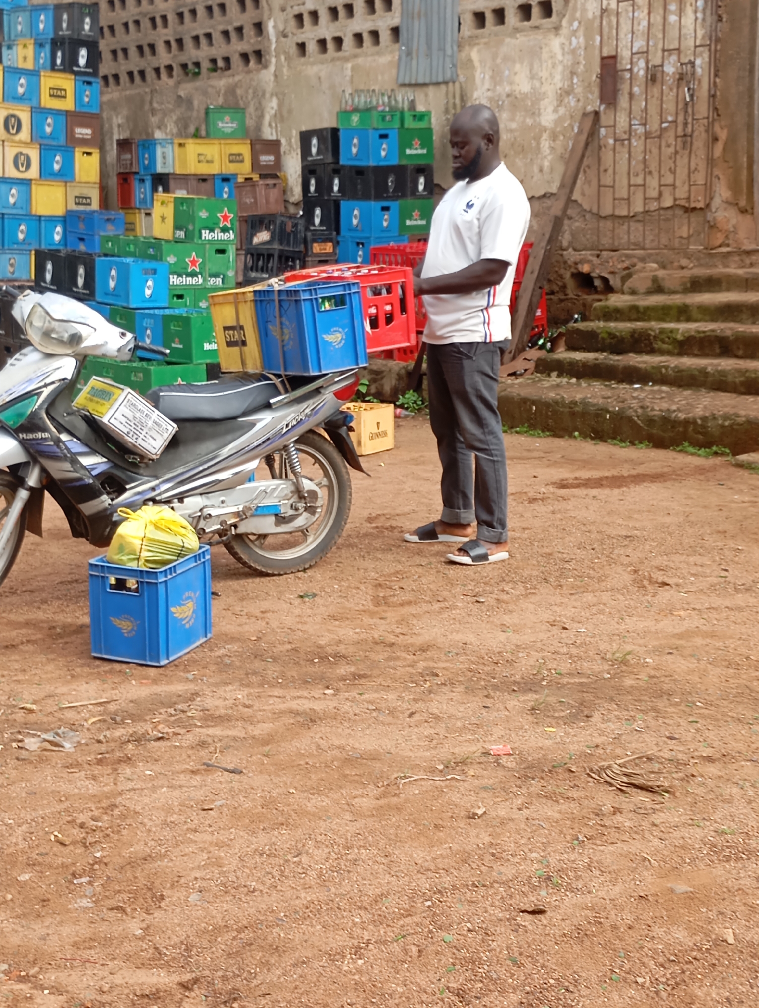 In Shendam, Nigeria, a middle-aged Black man wearing a white t-shirt and dark pants stands in a dusty, outdoor area piled high with plastic crates, many of which are red, blue, and yellow, and appear to be used for carrying beverages. He is looking down at the crates. A silver and black motorcycle is parked to his left, with a yellow plastic bag and a cardboard box on its seat. The background shows a brick wall with patterned openings and a set of concrete steps. The ground is covered in reddish-brown dirt, scattered with debris.