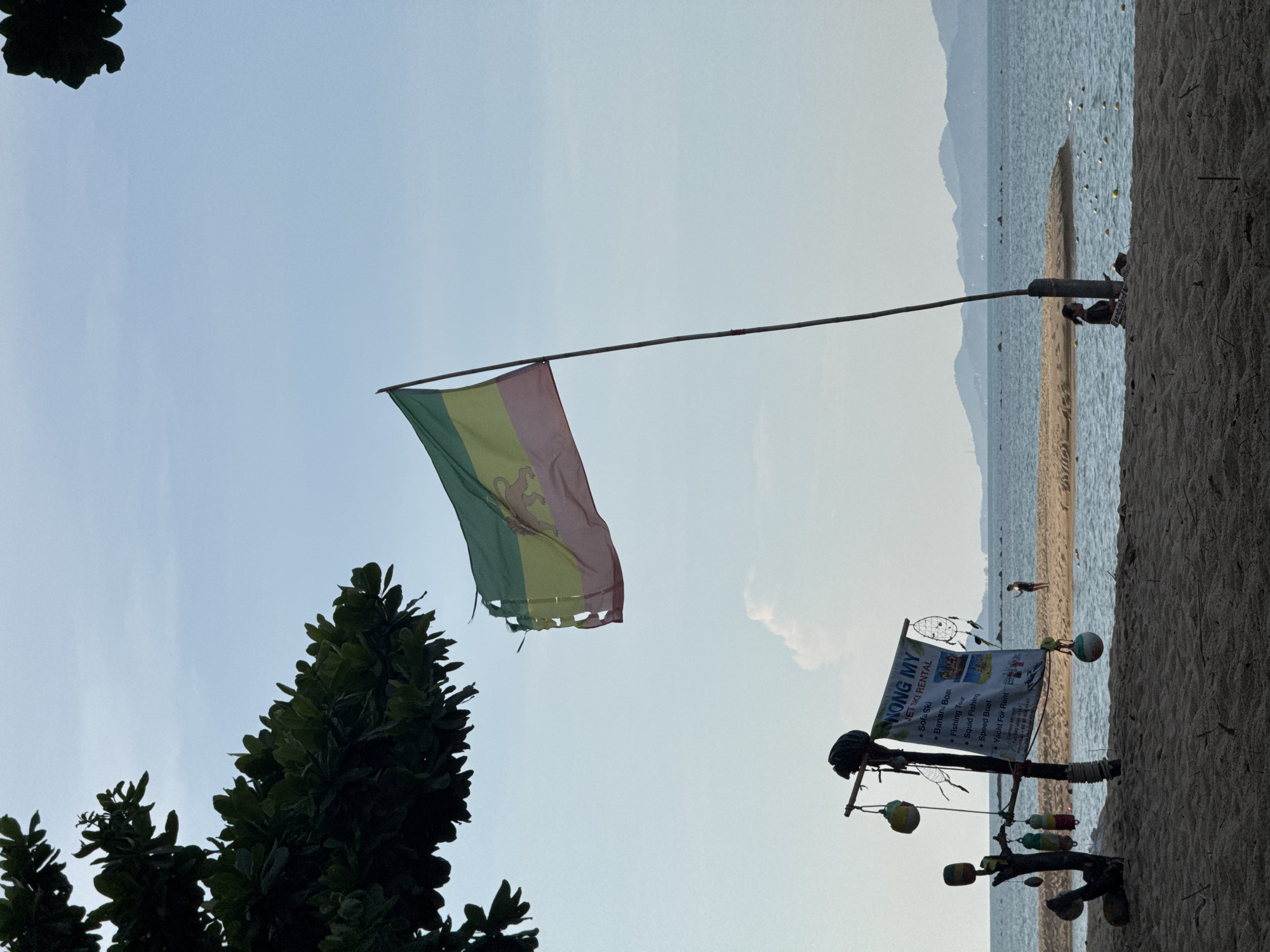 A wide-angle, outdoor, eye-level shot captures a beach scene in the Ko Samui District, Thailand, under a pale blue sky with wispy clouds. In the foreground, dark sand covers the beach. To the left, a dark green tree branch with large leaves extends from the top left corner of the frame. A flag, with horizontal bands of green, yellow, and pink, and a lion motif on the yellow band, is attached to a tall, thin pole on the right side of the image. The flag appears worn and torn.
On the left side of the foreground, a pole with several decorative items hangs from it, including dream catchers and colorful hanging objects. A sign board is attached to this pole, listing "NONG MY JET SKI RENTAL" and various activities like "Sofa Ski," "Banana Boat," "Fishing Tour," "Squid Fishing," "Speed Boat," and "Yacht For Rent." Below the services, contact information and a social media handle are listed.
Further back, a sandbar extends into the blue water, where a person is seen standing in silhouette near the far end of the sandbar. In the middle ground, the ocean stretches to the horizon, with faint mountains visible in the distance. A few small, yellow buoys are scattered in the water. In the right foreground, a person sits on the sand near the base of the flag pole, looking down at a phone. The overall atmosphere is serene and relaxed.