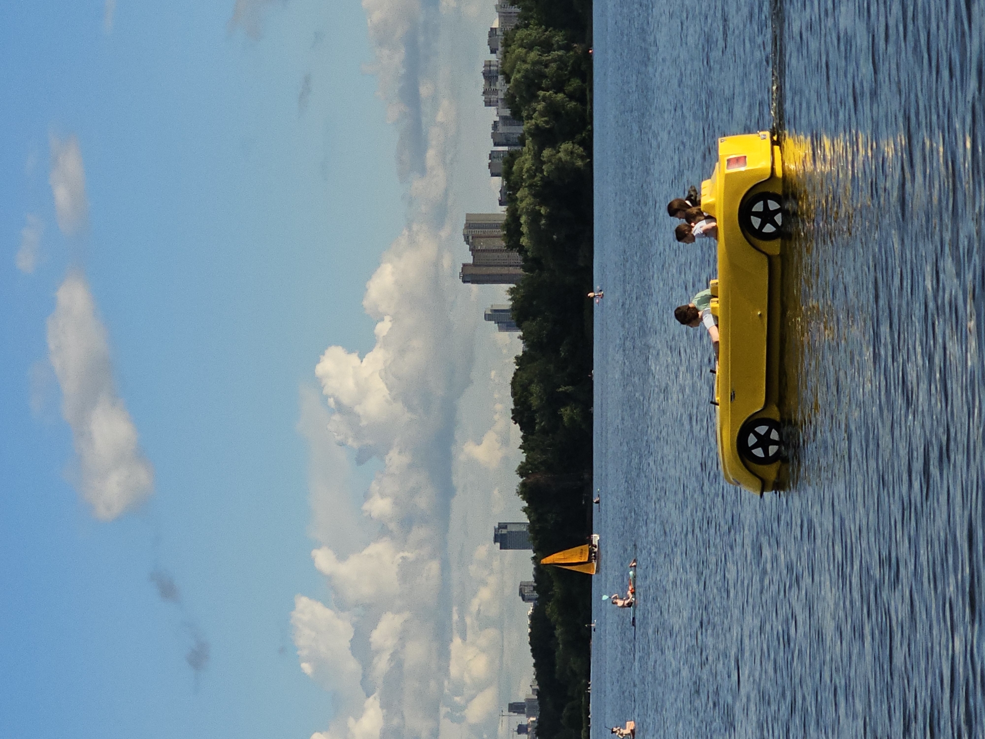 In Khimki, Russia, on a sunny day with scattered clouds in a blue sky, a yellow pedal boat shaped like a car navigates a calm body of water. The boat, with two children and possibly an adult inside, is in the foreground, leaving a slight wake. In the middle ground, several people are engaged in water activities, including individuals on paddleboards and a small sailboat with an orange sail. The background features a dense line of green trees and a cityscape with several tall buildings. The overall scene depicts a recreational day at a lake or river in an urban setting.