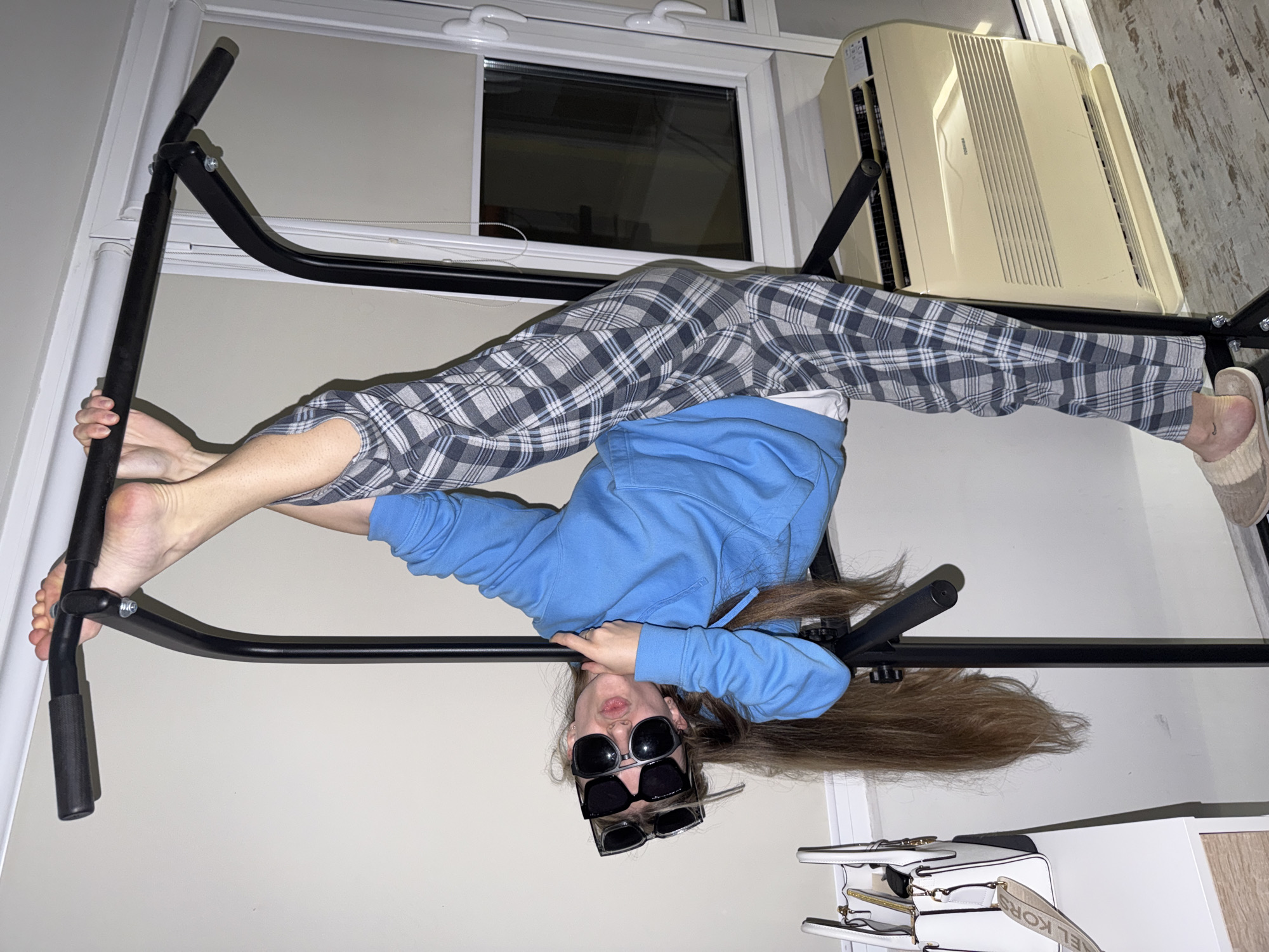 A person is performing a split on a doorway pull-up bar. The person is wearing plaid pants, a blue hooded sweatshirt, and two pairs of sunglasses. The location is an indoor setting. The city is not discernable.