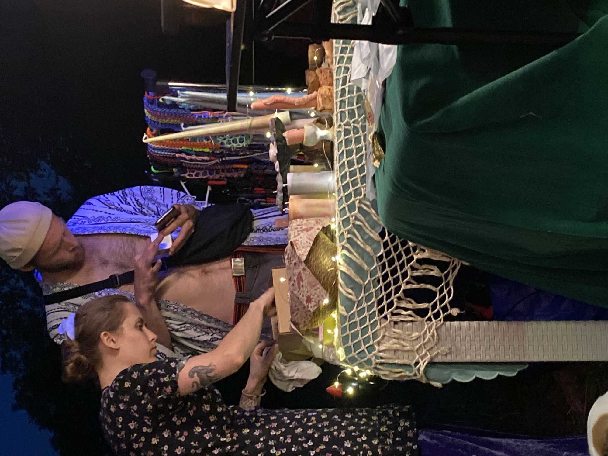 This is a medium shot, taken at night, of two individuals at a market stall in Asureti, Georgia. The person on the left, a female, wears a dark floral print dress and has her hair pulled up. She is leaning over a table, seemingly examining something within a small cardboard box. She has a tattoo of a black panther on her left arm. Beside her, a male wearing a light-colored beanie and an open, patterned robe is looking down at a mobile phone, holding it in his right hand. He has a light beard and is shirtless, revealing a metal belt buckle. The stall displays a variety of items, including strings of colorful braided bracelets, several tall candles, decorative objects, and what appear to be various trinkets. The table is covered with a patterned cloth and adorned with string lights. A green cloth drapes over a portion of the table in the foreground. The background is dark, suggesting an outdoor setting at night.