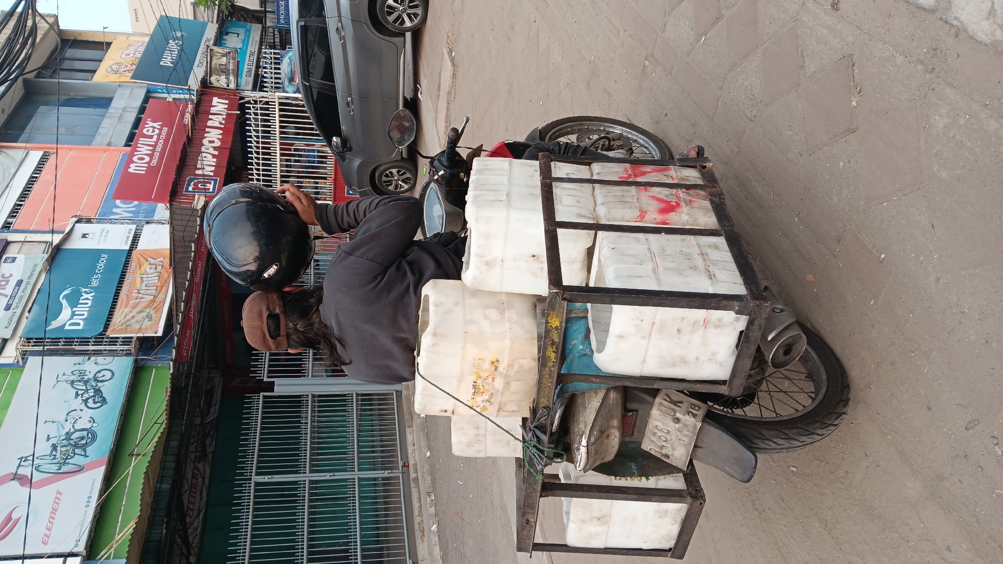 The image depicts a street scene in Tangerang, Indonesia, likely a commercial area given the presence of numerous storefronts and advertising banners. The foreground is dominated by a motorcycle with a large, overloaded cargo rack at the rear. The rack is filled with multiple white plastic containers, some stacked several high, suggesting a delivery or transport operation. A person, seen from behind, is seated on the motorcycle, holding a black helmet in their right hand and wearing a brown baseball cap. The motorcycle is parked on a paved street, with a grey car visible in the background. The storefronts behind the motorcycle display various brand advertisements, including "Dulux," "Vinilex," "Mowilex," and "Nippon Paint." The overall atmosphere suggests a typical urban environment in Indonesia, with a focus on commerce and daily transportation.