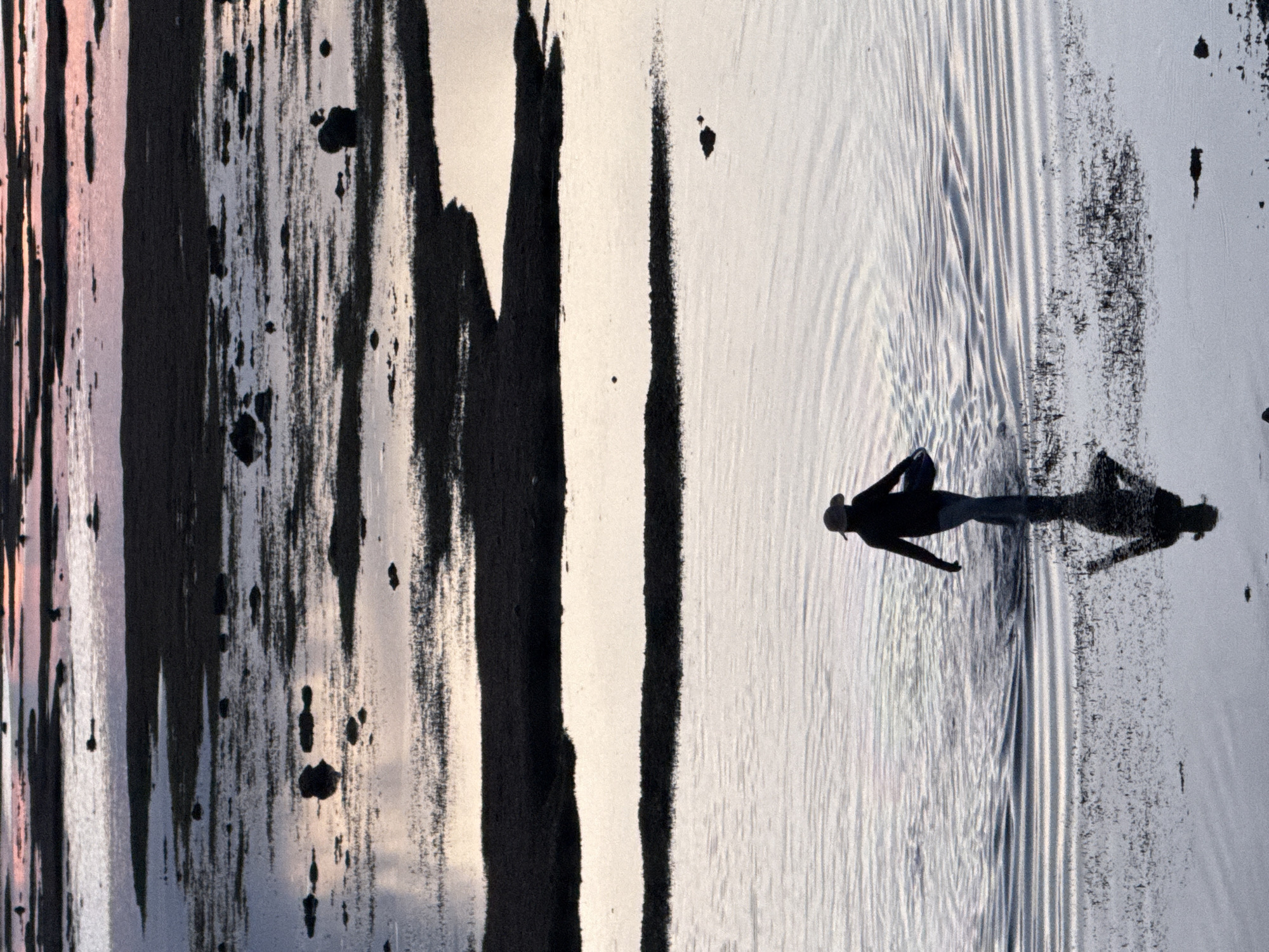 This is a vertical, abstract photograph featuring a solitary person silhouetted against a body of water. The image is divided into two distinct sections by what appears to be a large, dark, abstract brushstroke or a natural feature with a similar texture, running from top to bottom on the left side. This section is a mixture of dark, vertical strokes and splotches, with hints of pink and grey reflecting from a light source.
The right side of the image depicts a bright, shimmering surface, possibly water, with ripples and reflections creating a pattern of horizontal lines. A silhouetted figure is positioned in this water, facing left, appearing to be in motion. The figure is wearing what looks like a hat. A distinct shadow of the figure is cast on the water, extending to the right. The overall impression is that of a person walking or wading through shallow water at a time of day when the light is low, casting long shadows. The setting appears to be an outdoor environment, likely a beach or a shallow coastal area, given the presence of water and the low angle of the light suggesting either sunrise or sunset. The abstract elements on the left could be interpreted as natural rock formations, or perhaps intentional artistic elements added to the scene. There is no visible text in the image.