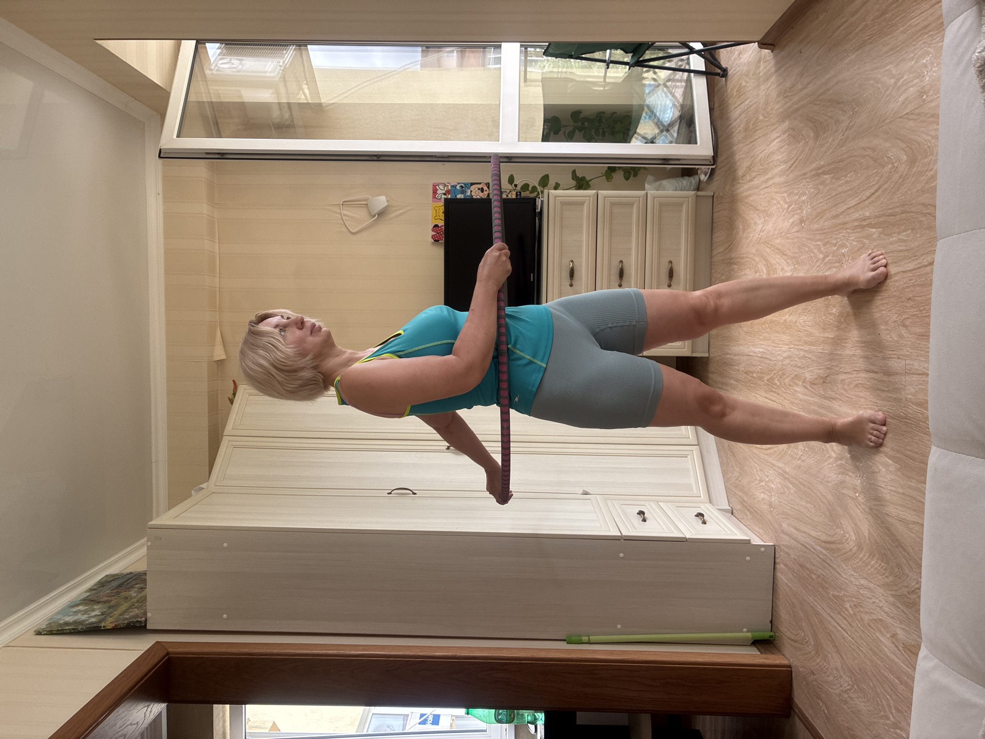 A Caucasian woman with blonde hair is standing in a room and is in the process of exercising with a hula hoop. She is wearing a teal tank top with a yellow trim and teal shorts. The woman is holding the hula hoop around her waist. The room appears to be a living room or bedroom, with a large wooden closet on the left side of the frame, a dresser with a television on top, and a balcony door on the right. The flooring is a light-colored wood laminate. The walls are covered in a light beige wallpaper with a vertical stripe pattern. There is a painting on the wall above the closet, and a white object hanging on the wall behind the television. The scene is well-lit, suggesting natural light from the balcony. The overall impression is of a domestic environment where an indoor workout is taking place.