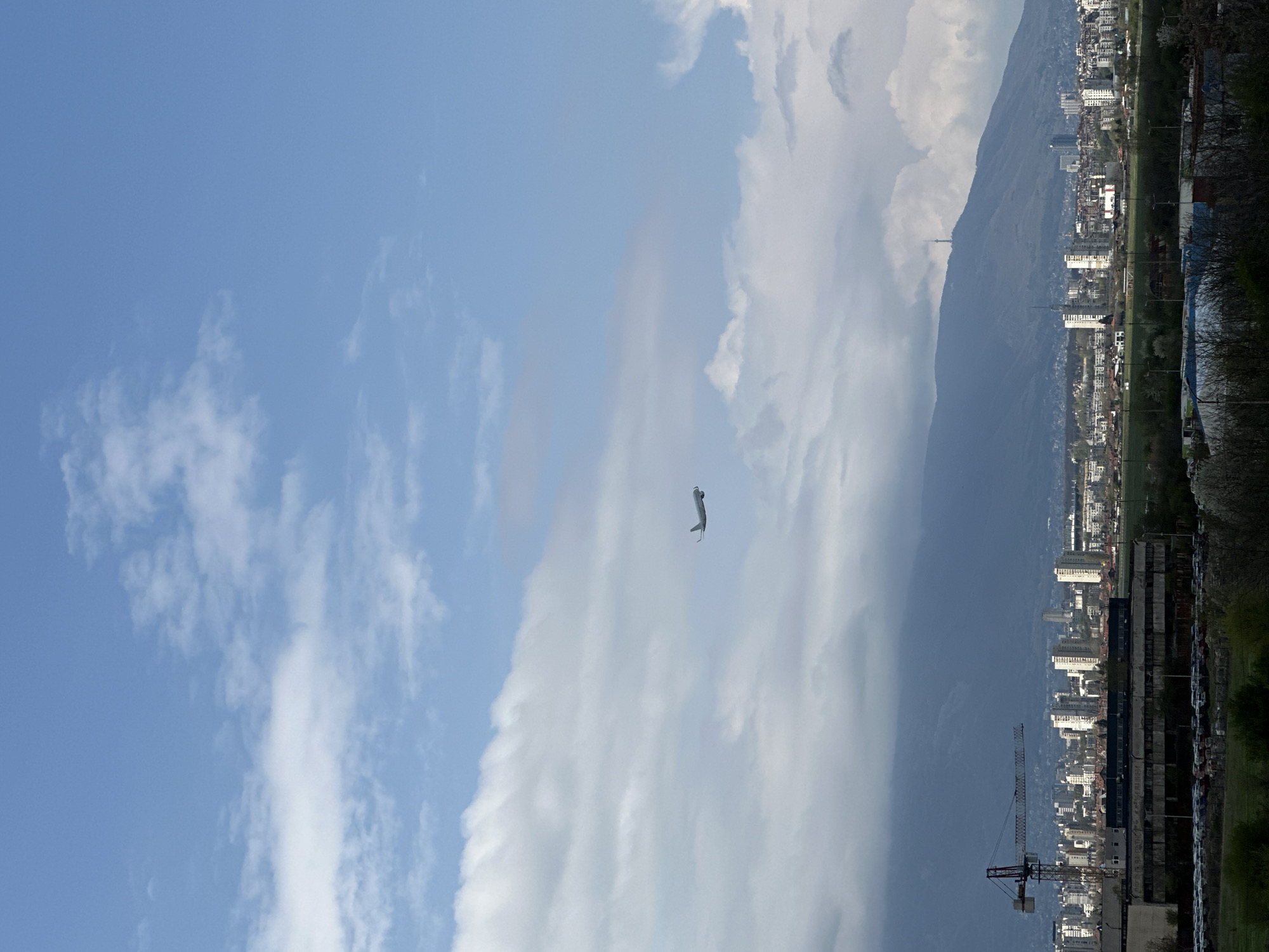 The image shows a daytime scene in Sofia, Bulgaria, with a focus on the sky. The sky is predominantly blue with patches of white clouds. In the foreground, a white, rigid paraglider is descending. In the background, there is a mountain range obscured by clouds. At the base of the mountain, a portion of the city of Sofia is visible, including buildings, a crane, trees, and vegetation.