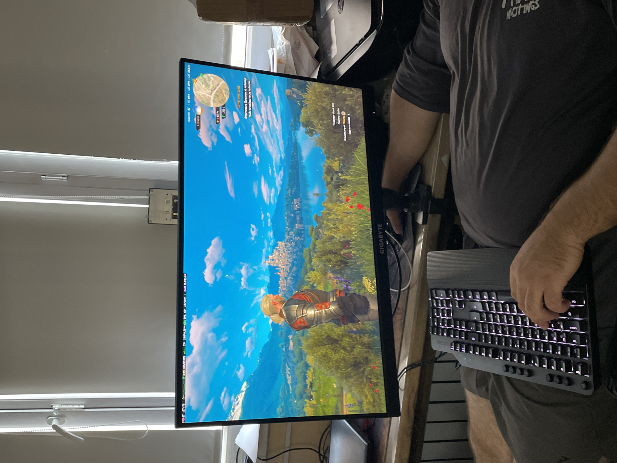 The image shows a person playing a video game on a computer. The game appears to be The Witcher 3: Wild Hunt, judging by the in-game graphics and HUD elements. The person is sitting in front of a large monitor, and a keyboard is visible on their lap. The room appears to be a home office or living space, with blinds on the window and some office supplies in the background. The overall scene is casual and relaxed. The image does not provide any information about the specific location in the city of Almaty, Kazakhstan.