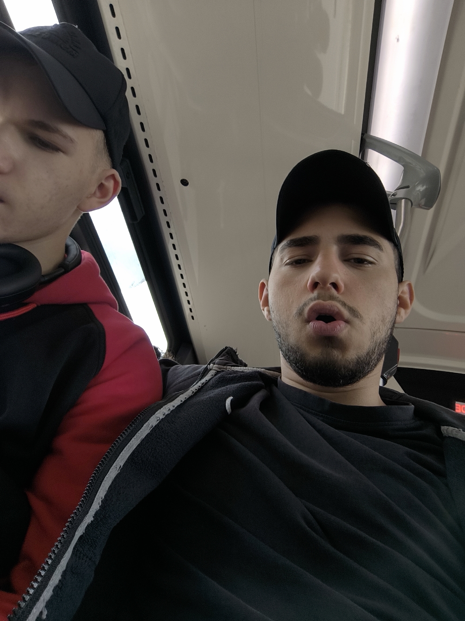 Two men are in a bus or train. The man in the foreground is wearing a black t-shirt, a black jacket with a white stripe, and a black baseball cap. He has a beard and mustache. His mouth is slightly open. Beside him, the other man wears a black and red hoodie and a black baseball cap. Headphones are around his neck. They sit in front of a window and the top of a grey handrail is visible. The location is most likely the city of Strasbourg in France.