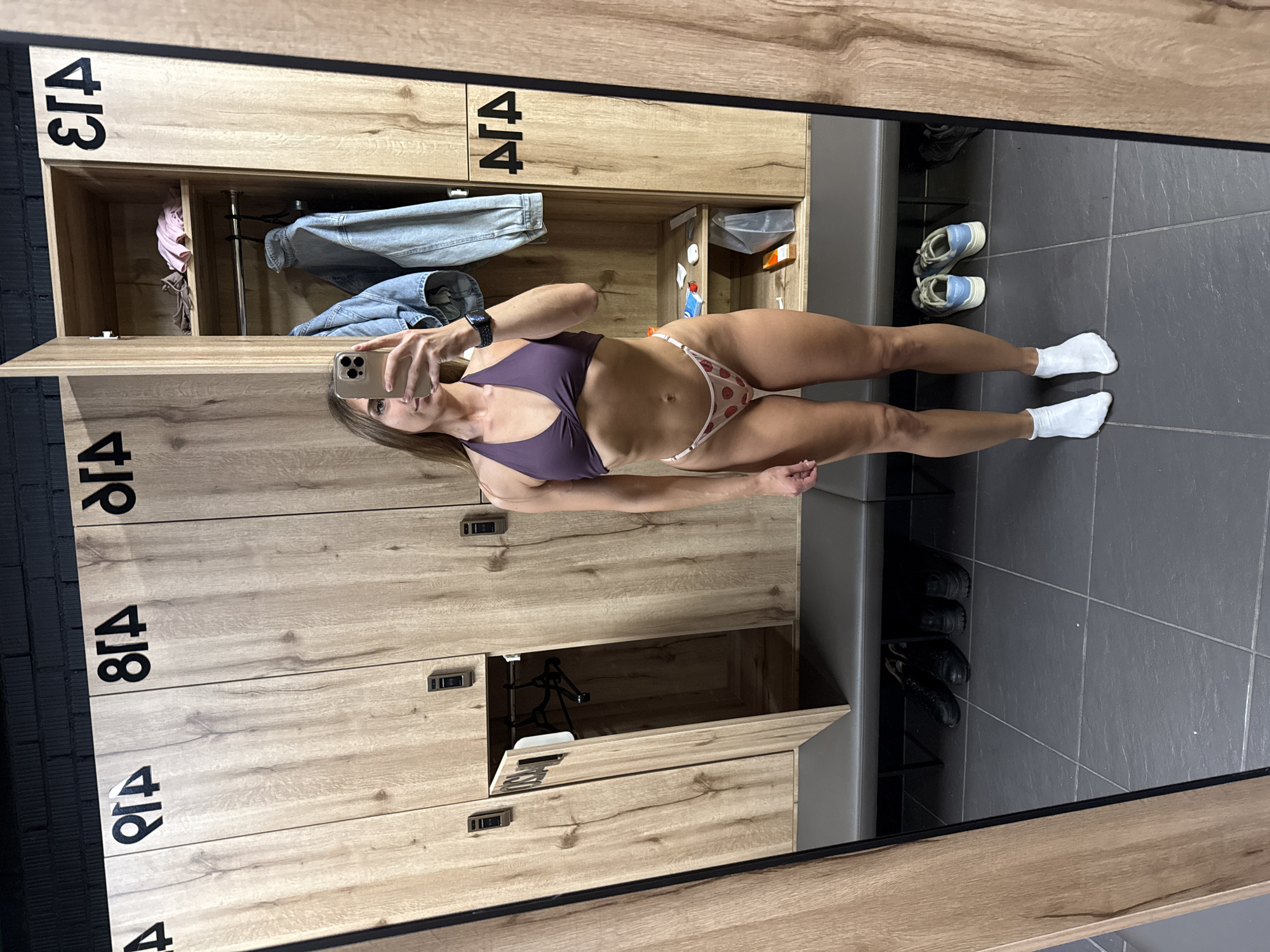A young woman with long, light brown hair is taking a mirror selfie in what appears to be a locker room. She is wearing a purple halter-neck bikini top and a white bikini bottom with a strawberry print. She has on white socks. In the background, there are wooden lockers, some of which are open, revealing clothing and shelves. There is also a bench and shoes on the floor. The locker numbers are visible and appear to be in a Cyrillic script. The scene is well-lit, and the woman is the primary subject of the image. The woman's face is partially obscured by her phone, but her body is clearly visible. The environment suggests a fitness facility or gym. There is no indication of the location being in Saint Petersburg, Russia.