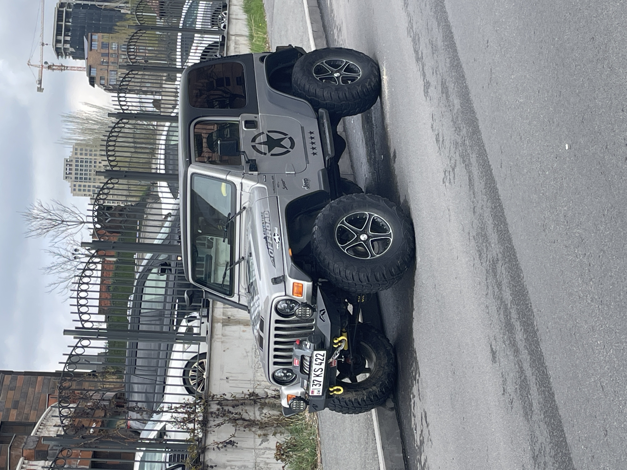 The image depicts a silver Jeep Wrangler parked on the side of a street in Yerevan, Armenia. The vehicle has oversized tires and a star graphic on the driver's side door. The license plate reads "37 KS 422." A black, wrought-iron fence runs along the adjacent property, behind which are visible buildings and a construction crane. The street is asphalt, showing some signs of wear. The sky is overcast.