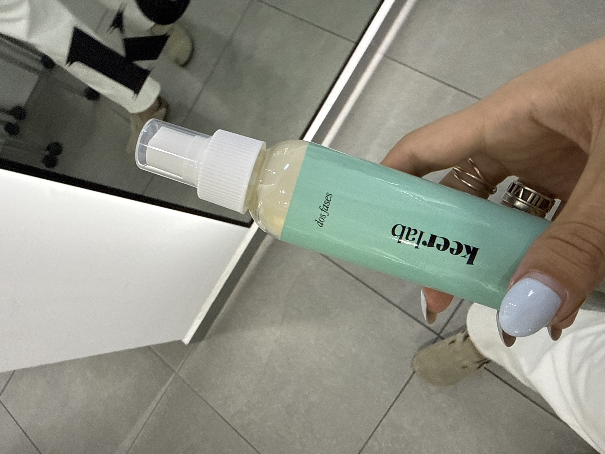 The image shows a hand holding a light green spray bottle with a white spray nozzle. The bottle is labeled "keerlab" in black text and "dos fases" in a smaller font. The hand has a light blue nail polish and wears multiple rings. The background includes tiled flooring and the reflection of a person in a mirror. The person in the reflection wears a black and white outfit. The photograph was taken indoors. No information indicates the city of San Bernardino Tlaxcalancingo, Mexico.