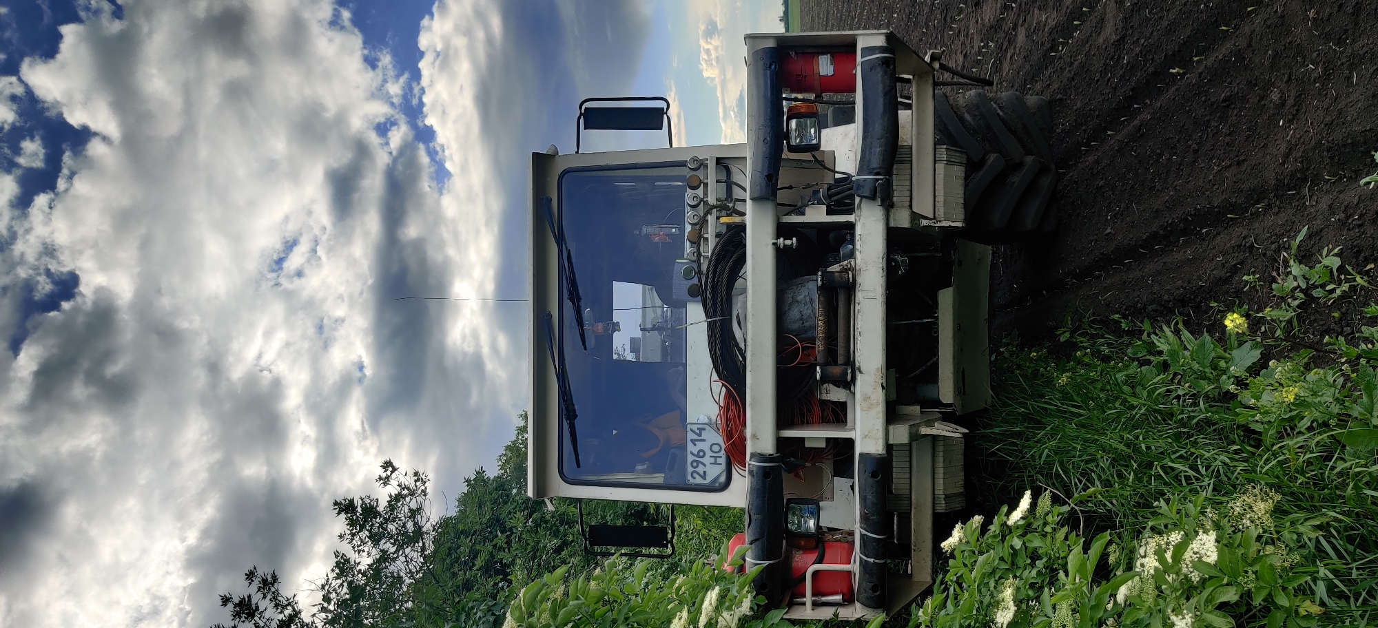 The image depicts an agricultural sprayer in a field near Kovalivs'ke, Ukraine. The machine is predominantly white, with a cabin, a set of large tires, and a visible license plate reading "29614 HO". A red fire extinguisher is mounted on the sprayer's frame. The sprayer is parked adjacent to a field with dark soil, and the foreground is filled with green vegetation and small white flowers. Above the sprayer, the sky is partly cloudy.
