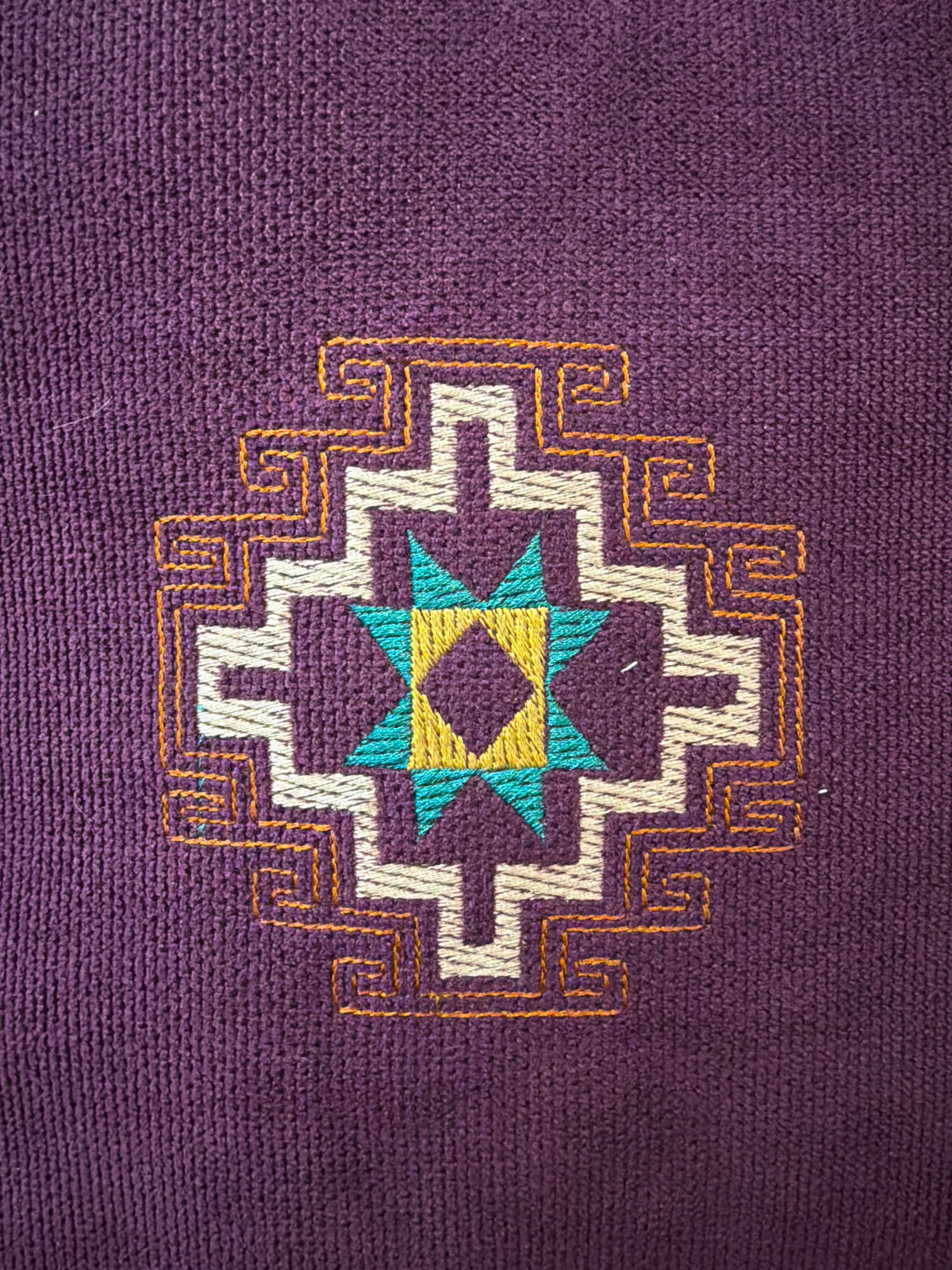 The image is a close-up, overhead view of an embroidered geometric design on a dark purple, textured fabric. The design is symmetrical and features a central eight-pointed star formed by alternating yellow and teal triangles. This star is enclosed within a diamond shape, also in yellow. Surrounding the central star is a complex, layered geometric pattern in cream and orange. The cream portions form stepped, angular shapes, creating a sense of depth. The orange embroidery outlines some of these shapes and forms intricate, spiral-like details in the corners. The fabric itself has a visible weave, suggesting it could be a textile such as a sweater or a tapestry. There are no people or other elements in the image, nor is there any context that would place it in Yerevan, Armenia.