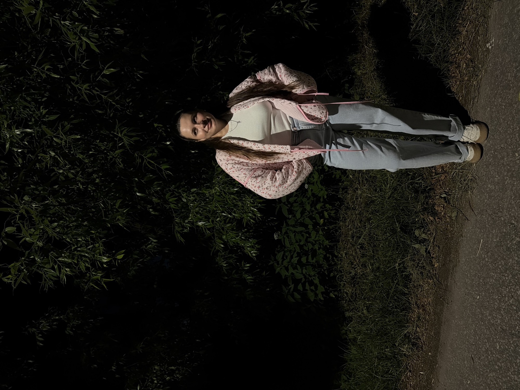A full-body shot captures a young woman standing outdoors at night, in what appears to be a park or wooded area near an asphalt path. She is facing the camera with her hands on her hips, looking slightly to her right. The woman has long, dark hair pulled back and is wearing a white ribbed top, light blue jeans, and light-colored sneakers. She is also wearing a pink quilted jacket with a floral pattern and white trim. Behind her, a dense dark green foliage forms a natural backdrop, with some lighter green bushes visible in the mid-ground. The ground in front of her is covered with grass and debris, transitioning to the dark asphalt path on the left. The lighting suggests an artificial light source is illuminating the scene from the front. The location is specified as Красный Маяк, Russia.