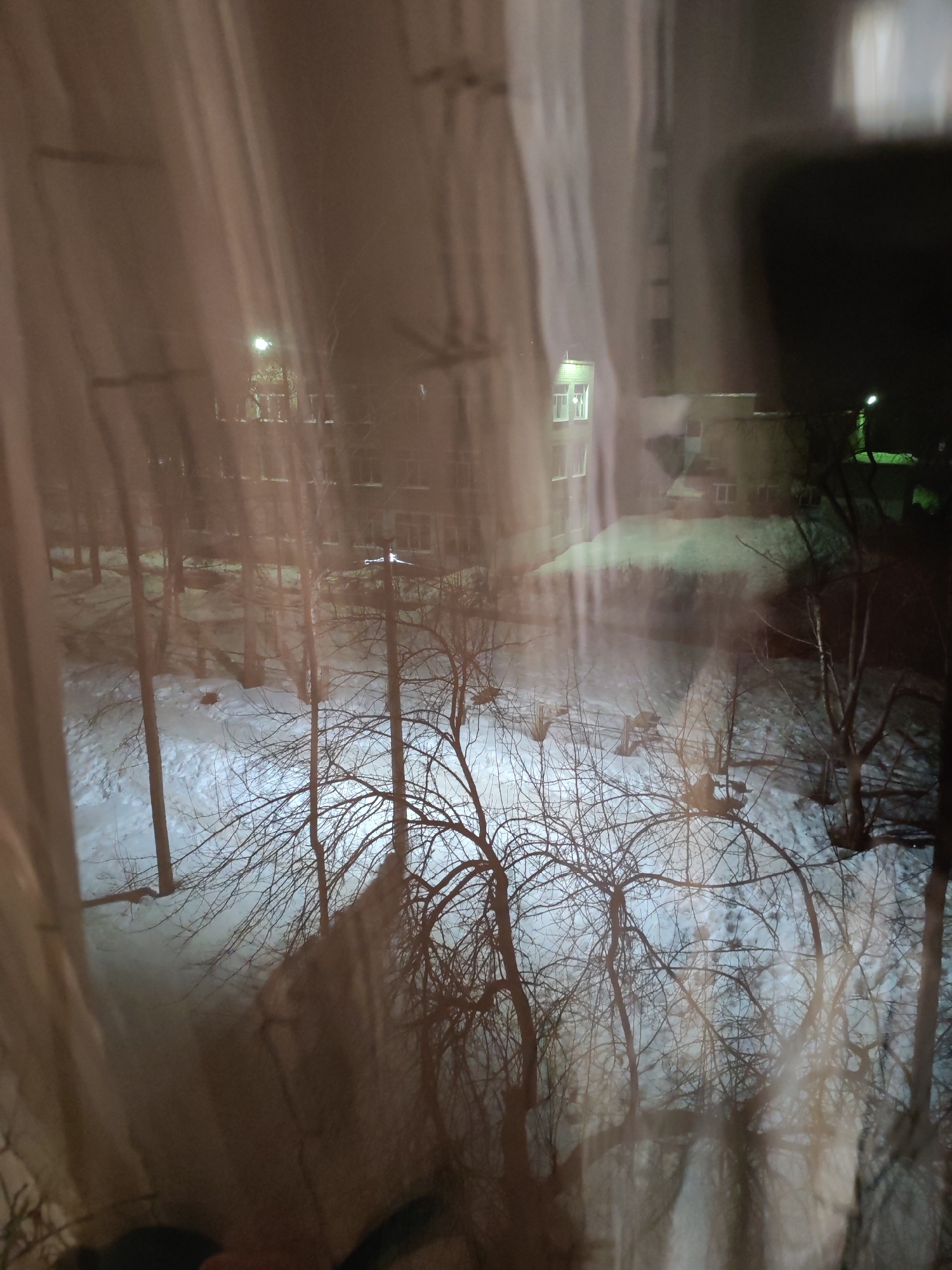 The image presents a nocturnal winter scene in the city of Chaikovsky, Russia, viewed from an interior perspective through a window. Light-toned curtains partially frame and obscure the left and right edges of the view, with reflections on the window glass creating a diffused effect. The ground outside is covered in white snow, traversed by numerous bare deciduous trees with visible branches. Artificial light sources illuminate sections of the snow-covered ground and tree canopies. A prominent streetlamp is visible above the tree line in the mid-ground. In the background, multi-story buildings are present, one on the left featuring several brightly lit rectangular windows. A distinct green light source is visible towards the far right of the scene. No individuals or specific actions are discernible within the frame.