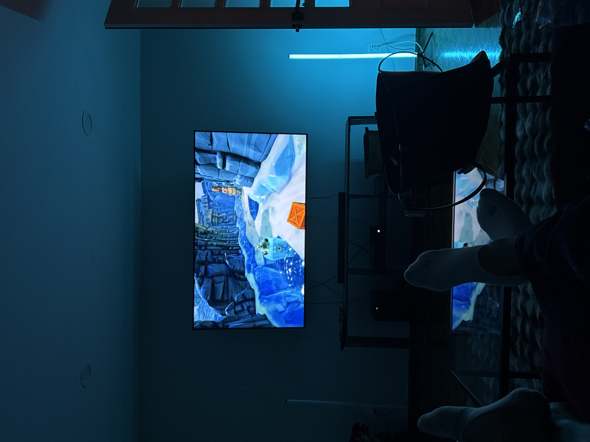 The image shows an interior scene dominated by a blue ambient light. A wall-mounted television displays a video game scene featuring a blue and white, icy landscape. Below the television is an entertainment center with shelves and devices. In the foreground, a person's legs and feet, clad in white socks, are visible. The overall scene evokes a sense of relaxed leisure. Location cannot be determined.