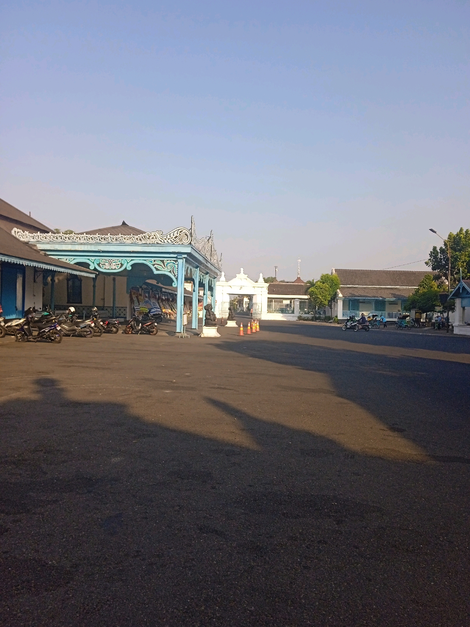 This is a landscape photograph taken on an overcast day in front of a traditional building complex. The building features a light blue ornate veranda with intricate carvings, supported by pillars. To the left of the veranda, a section of the building is visible with blue walls and white trim. Parked in front of the veranda are several motorcycles, and a shadow of a person can be seen on the paved ground. In the background, there is a white archway, more buildings with dark tiled roofs, trees, and a street lamp. A few more motorcycles and a rickshaw are visible further down the road. The overall scene appears to be in a historical or cultural district, likely a public space in Surakarta, Indonesia.