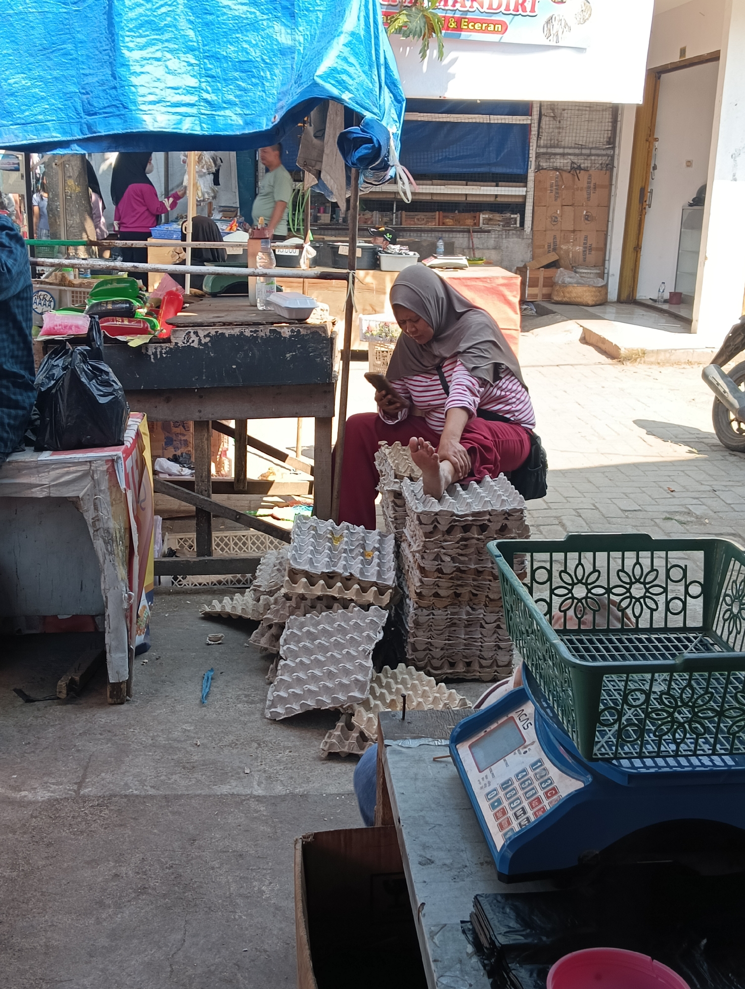 A woman wearing a hijab and a striped shirt is seated on a stack of egg cartons, holding a mobile phone. She is in an outdoor market setting in Katapang, Indonesia, indicated by a banner that reads "PASAR MANDIRI & ECERAN." The ground is paved with concrete. In front of her is a blue digital scale with a green plastic basket on top, and to her left, a black plastic bag is placed on a table. In the background, a blue tarp is stretched overhead, providing shade. Other market stalls and people are visible in the distance. The scene appears to be during the daytime, with sunlight casting shadows.