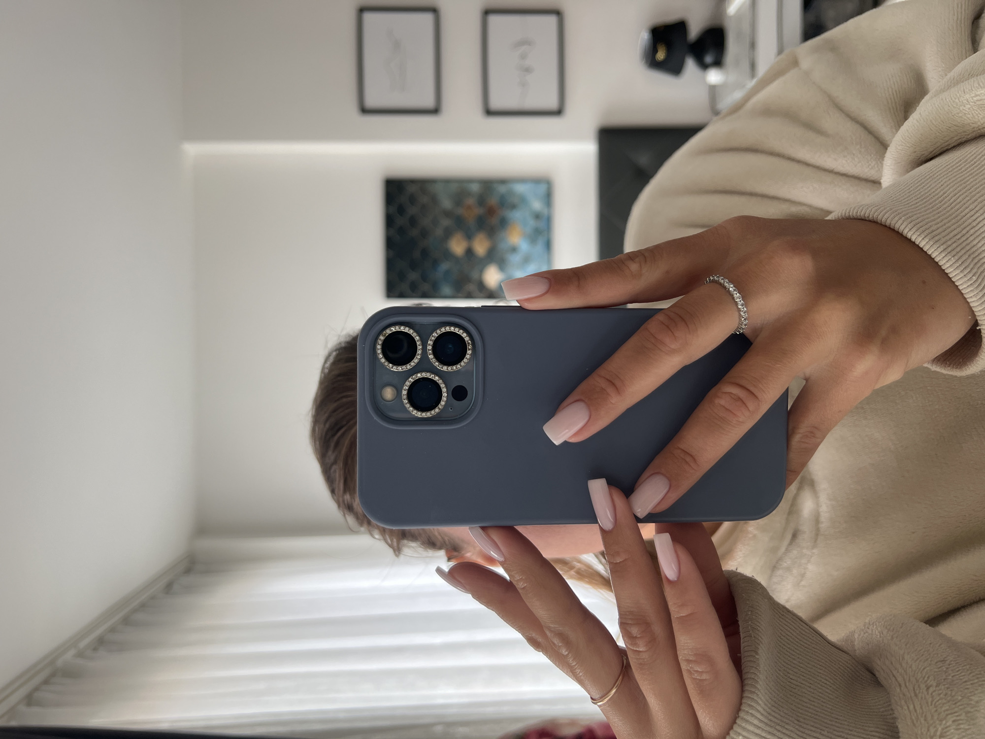 The image shows a person holding a blue phone, likely taking a selfie in a mirror. The background suggests a modern interior with framed artwork and decorative items. The person is wearing a beige-colored sweater, and the phone has a jeweled camera cover. It is not possible to infer the location in Budva, Montenegro or any other location from the provided image.