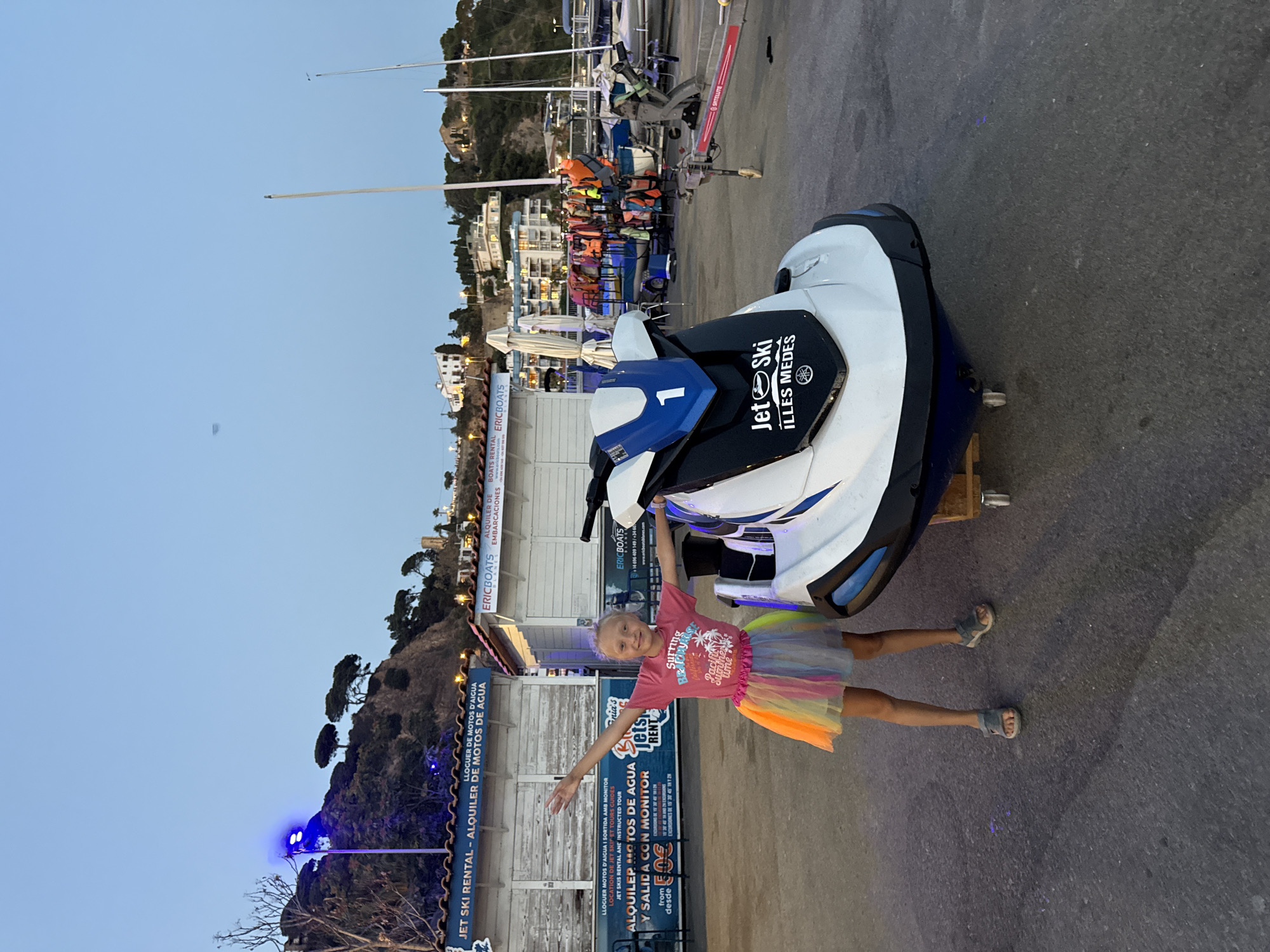 A young blonde girl wearing a pink t-shirt and a colorful tutu stands with her arms outstretched next to a white and blue jet ski. The jet ski has the number "1" on its front and "Jet Ski Illes Medes" written on its side. Behind them are several white rental booths with blue awnings. The booths have signs advertising jet ski rentals and boat tours. In the background, there are hills with trees and buildings, a harbor with sailboats, and a clear sky at dusk. The scene appears to be in the city of Blanes, Spain.