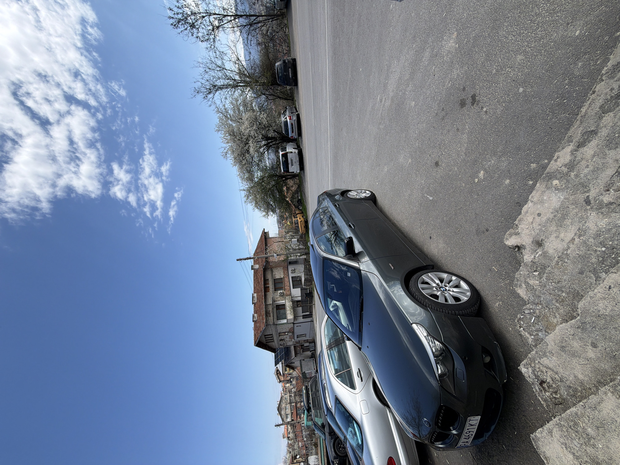 A medium-angle shot depicts a gray BMW sedan parked on an asphalt street in Sveti Konstantin, Bulgaria. The vehicle is adjacent to a concrete curb and other parked cars, including a silver sedan. Buildings with tiled roofs are visible in the background, along with trees and a blue sky with scattered clouds.