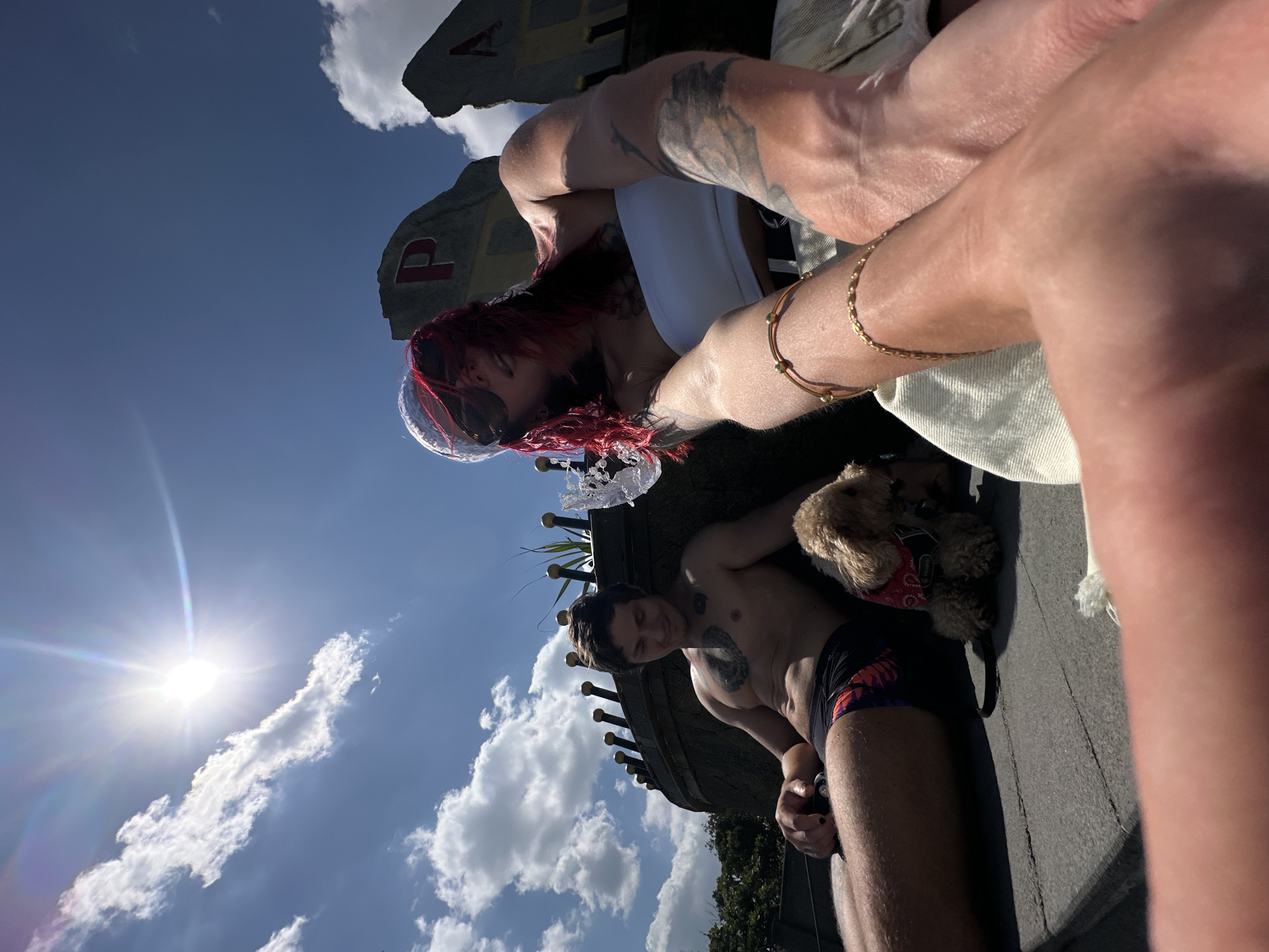 The image depicts an outdoor leisure scene in Mengwi, Indonesia, captured under a bright blue sky with scattered white cumulus clouds. A prominent sun flare originates from the upper left quadrant of the sky.
In the foreground, on the right side, a person with bright red hair, wearing a white headscarf and dark sunglasses, is positioned. They are clad in a white strapless bandeau top and light-colored bottoms, with multiple tattoos visible on both arms. Their right wrist displays a gold chain bracelet and another simpler gold bracelet. Their arms are extended towards the camera, suggesting a close-up perspective or that they are holding the camera, with their legs also angled forward.
To the left and slightly behind the foreground subject, a male figure is seated, bare-chested, showcasing a toned physique. He has several chest tattoos, including a large, dark, intricate design on his upper left chest and a circular design on his right chest. He is wearing dark swim trunks with a colorful, abstract pattern. His left thumbnail is painted with dark nail polish. He is looking downwards with a relaxed expression, his left hand resting on his leg and holding a small, dark, indistinct object.
Between the two individuals, a small, light brown, curly-haired dog, resembling a toy poodle, sits calmly. The dog is wearing a red bandana with white spots around its neck and a black harness with an attached leash.
In the background, two large, rock-like structures with bold red letters painted on them are partially visible on the right. The left structure clearly shows the letter "P," while the right shows a partial "A." Behind the male figure, a dark, low-lying structure, possibly a wall or the railing of a boat, is adorned with a series of golden, sword-shaped finials along its top edge. A splash of water is visible immediately to the right of this structure, indicating a water feature. Green foliage is discernible on the far left behind the dark structure. The subjects are resting on a flat, dark grey, textured surface.