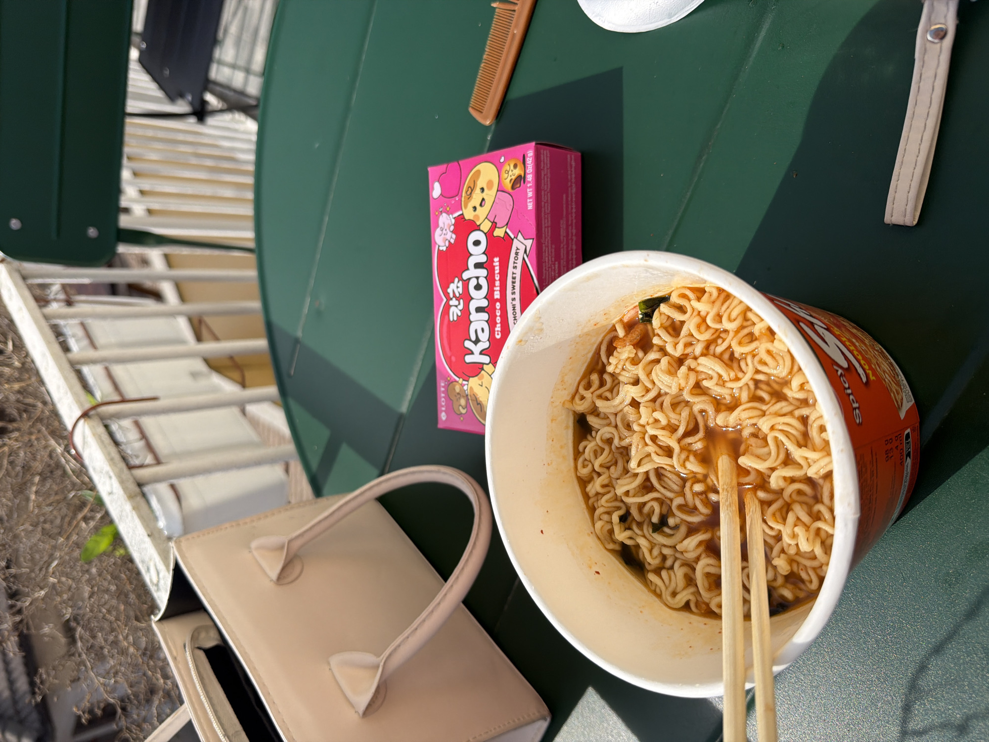 The scene depicts a close-up view of a green metal outdoor table on what appears to be a balcony in Veliko Tarnovo, Bulgaria, under bright daylight. A white cup of instant ramen noodles with wooden chopsticks submerged in the broth occupies the foreground. To the right of the ramen cup, a pink box of "Kancho Choco Biscuit" is visible, along with a small brown comb. A partial white plastic spoon head is present near the bottom right edge. A beige handbag with two handles rests on the table to the left of the ramen cup. Below the ramen cup, a small beige strap with a metallic snap is visible. In the background, a white metal railing and a partially visible dark green metal chair are present. Beyond the railing, out-of-focus bare tree branches and some green foliage are visible, suggesting an outdoor residential or urban environment. Strong sunlight casts distinct shadows on the table surface. No people are visible.