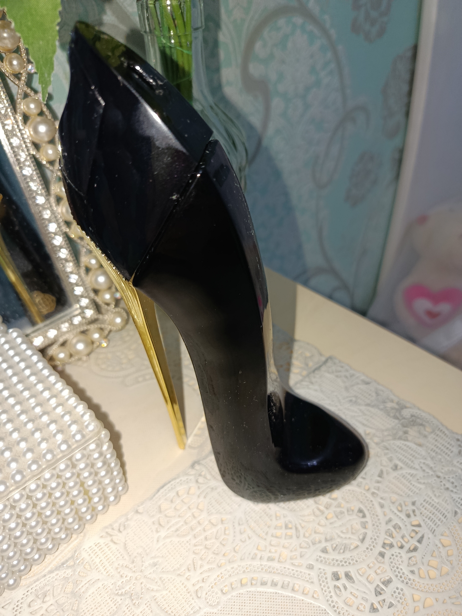 A close-up, slightly angled, indoor shot focuses on a black, high-heeled stiletto perfume bottle positioned on a textured white surface, possibly lace or a doily. The bottle is a deep, glossy black and features a striking gold stiletto heel. To the left of the bottle, a decorative object with an ornate, pearl-encrusted frame and pearls is visible, reflecting light. Further to the left, a portion of a green object, possibly a plant or vase, is seen. The background consists of a light blue wallpaper with a light gray damask pattern. In the far background, a blurred image of a white teddy bear with a pink heart detail can be discerned. The overall setting appears to be a dressing table or vanity area, suggesting a personal or decorative display. The image is taken in Tashkent, Uzbekistan.