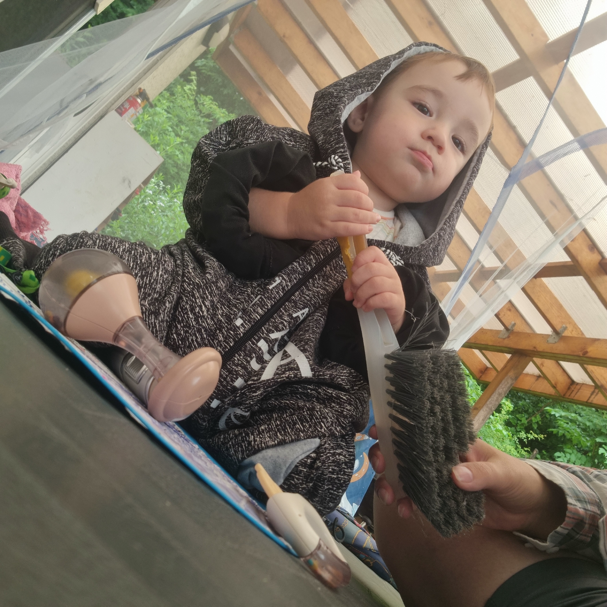 A low-angle, outdoor, medium shot captures a toddler boy sitting in a shelter with a wooden roof and clear plastic sheeting. The boy, facing left and looking toward the camera with a pout, wears a hooded black and grey patterned sweatshirt. He holds a yellow object with his right hand. In front of him on the ground are a pink cloth, a clear and tan baby rattle, and a white and clear baby nasal aspirator. To the right, a hand, possibly an adult's, holding a dark-bristled brush, is partially visible. The background shows green foliage. The scene is likely located in Dunnae-myeon, South Korea, based on the provided metadata.