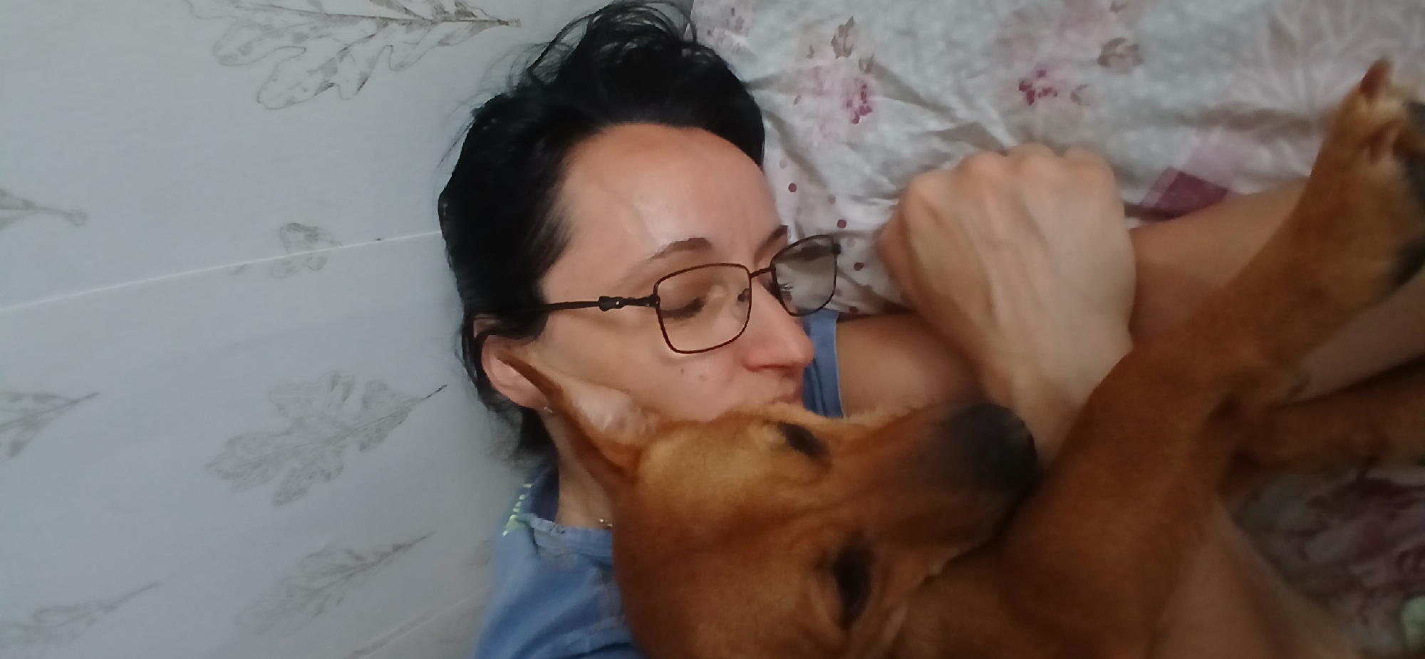 A woman with dark hair and glasses is lying down and cuddling a brown dog. She is wearing a blue shirt, and the dog is resting its head on her shoulder. They are on a bed with floral-patterned sheets, and a wall with a leaf pattern is visible in the background. The image appears to be a personal photograph taken in a domestic setting, likely a bedroom in a residence in Blagoevgrad, Bulgaria.
